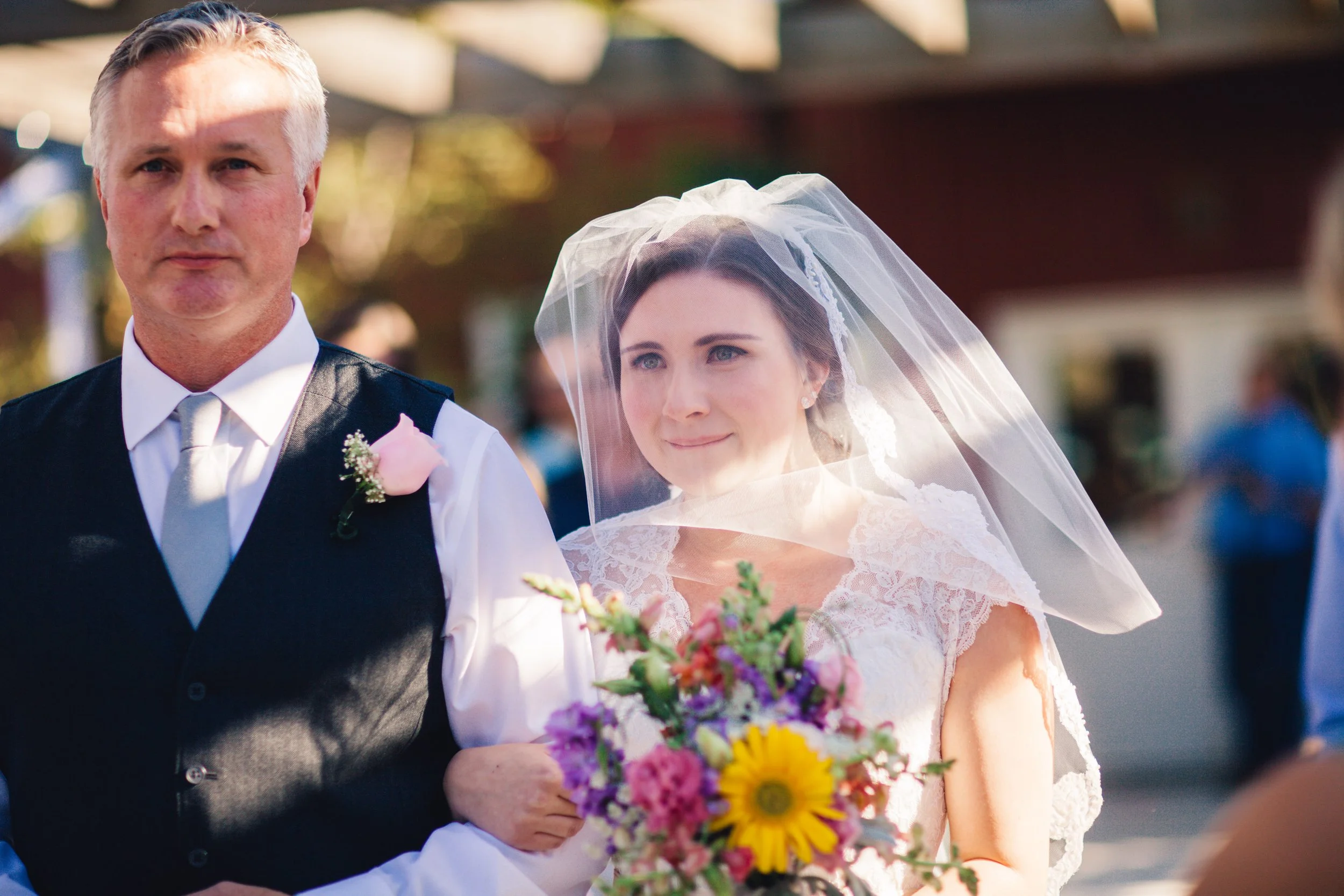 A bride with a veil holding a colorful bouquet, standing next to a man in formal attire during an outdoor wedding ceremony.