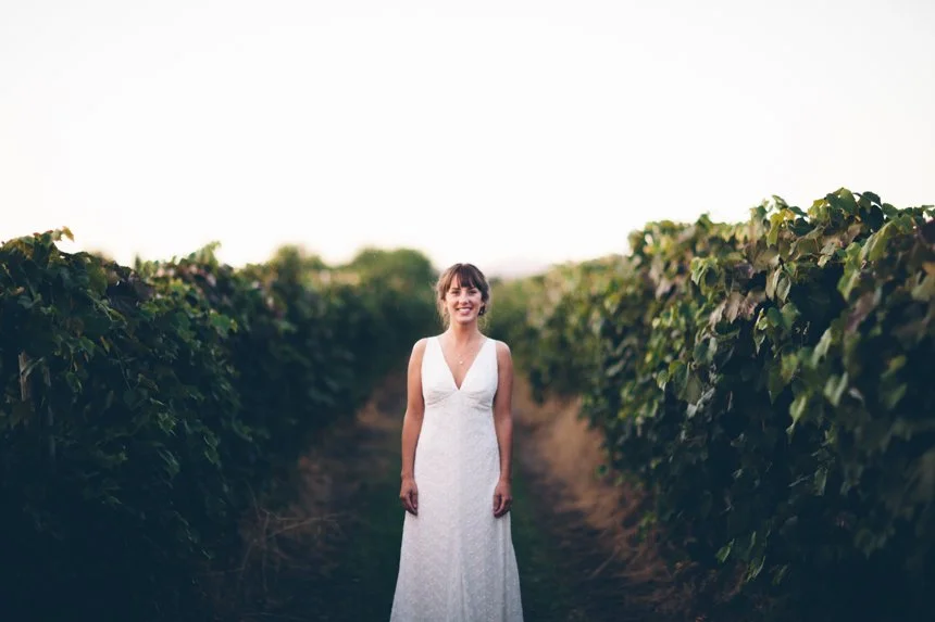 Woman in a white wedding dress standing between rows of green grapevines in a vineyard.