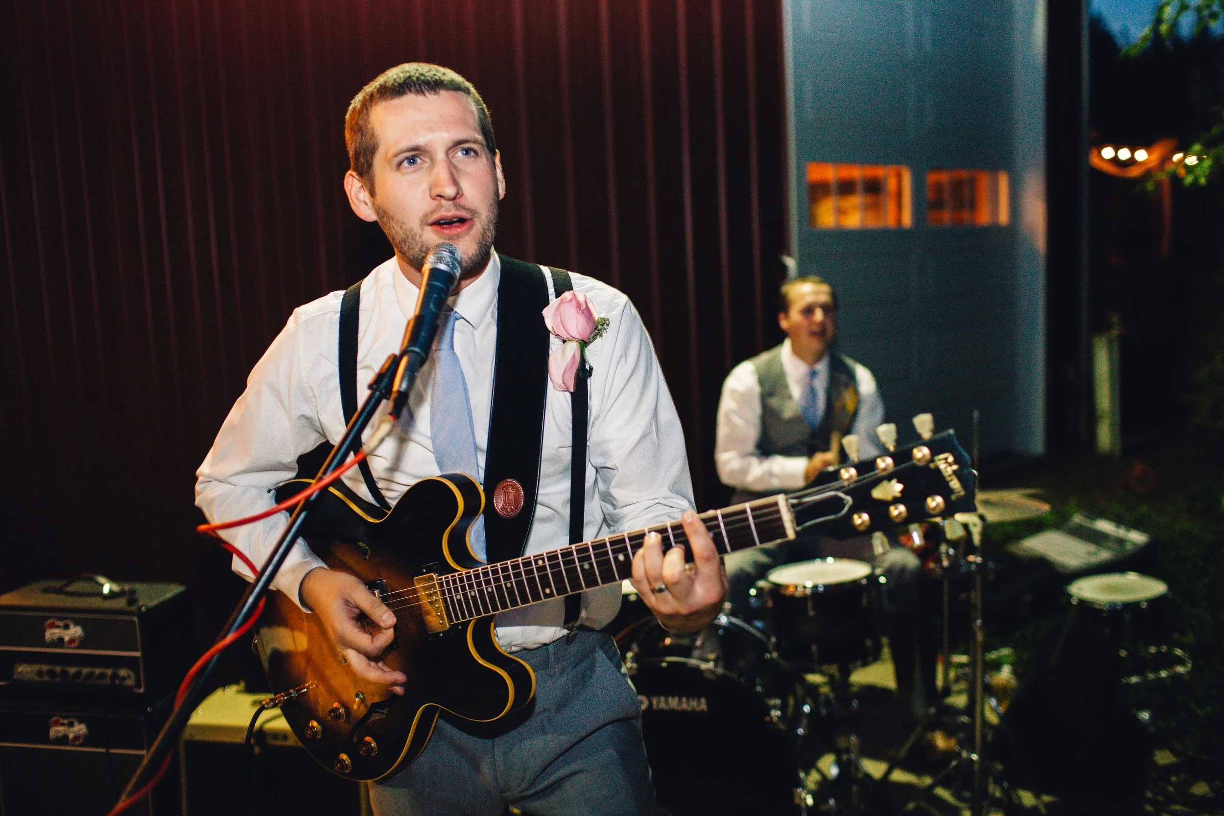 A man playing an electric guitar and singing into a microphone at an indoor event. A drummer is visible in the background, and both are dressed in formal attire with suspenders and a boutonniere.
