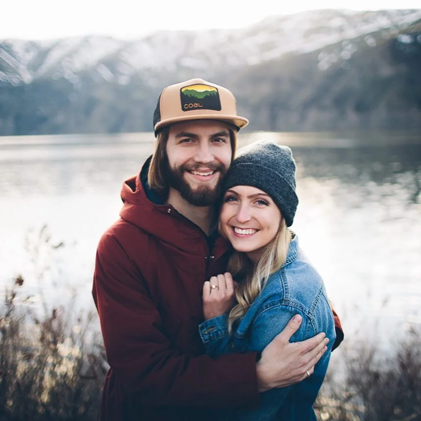 Smiling couple in outdoor setting by a lake with mountains in the background, hugging each other, woman wearing a gray beanie and denim jacket, man wearing a beige cap and red jacket.