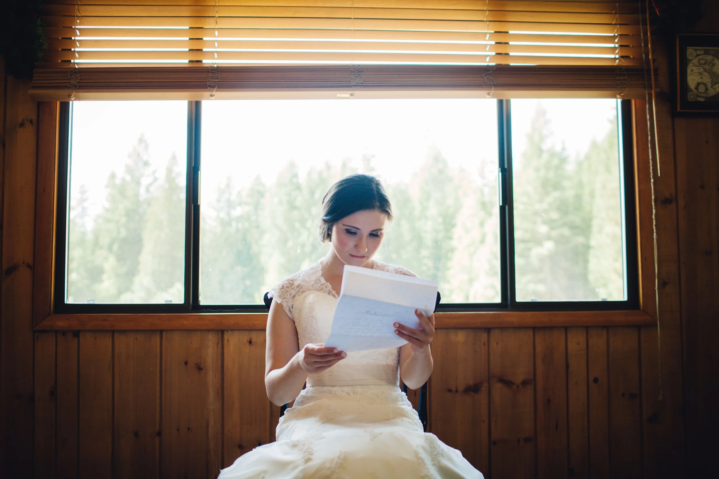 A bride in a white wedding dress sitting in front of a large window with wooden blinds, reading a letter or note.