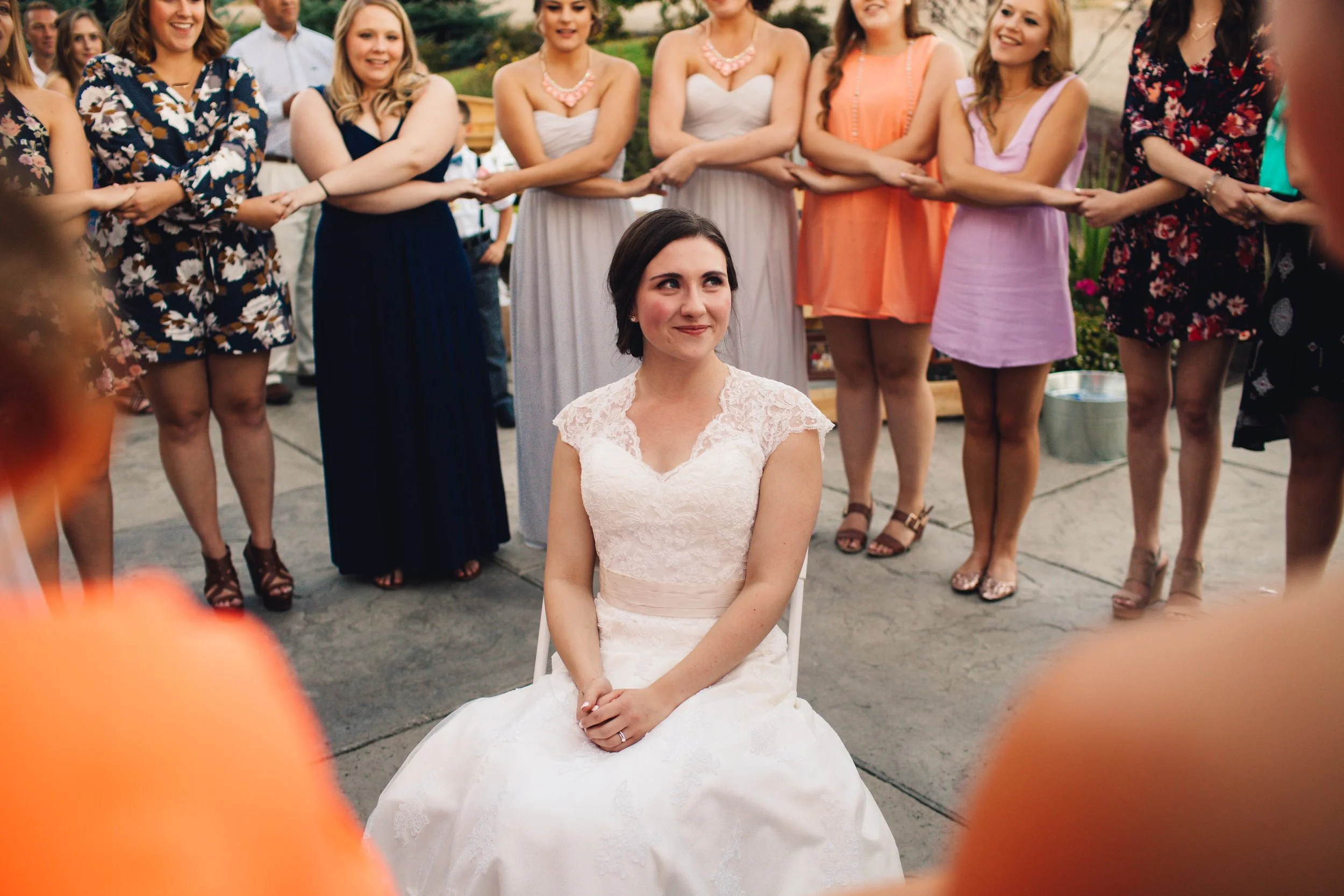 Bride seated outside surrounded by women in dresses at a wedding reception.