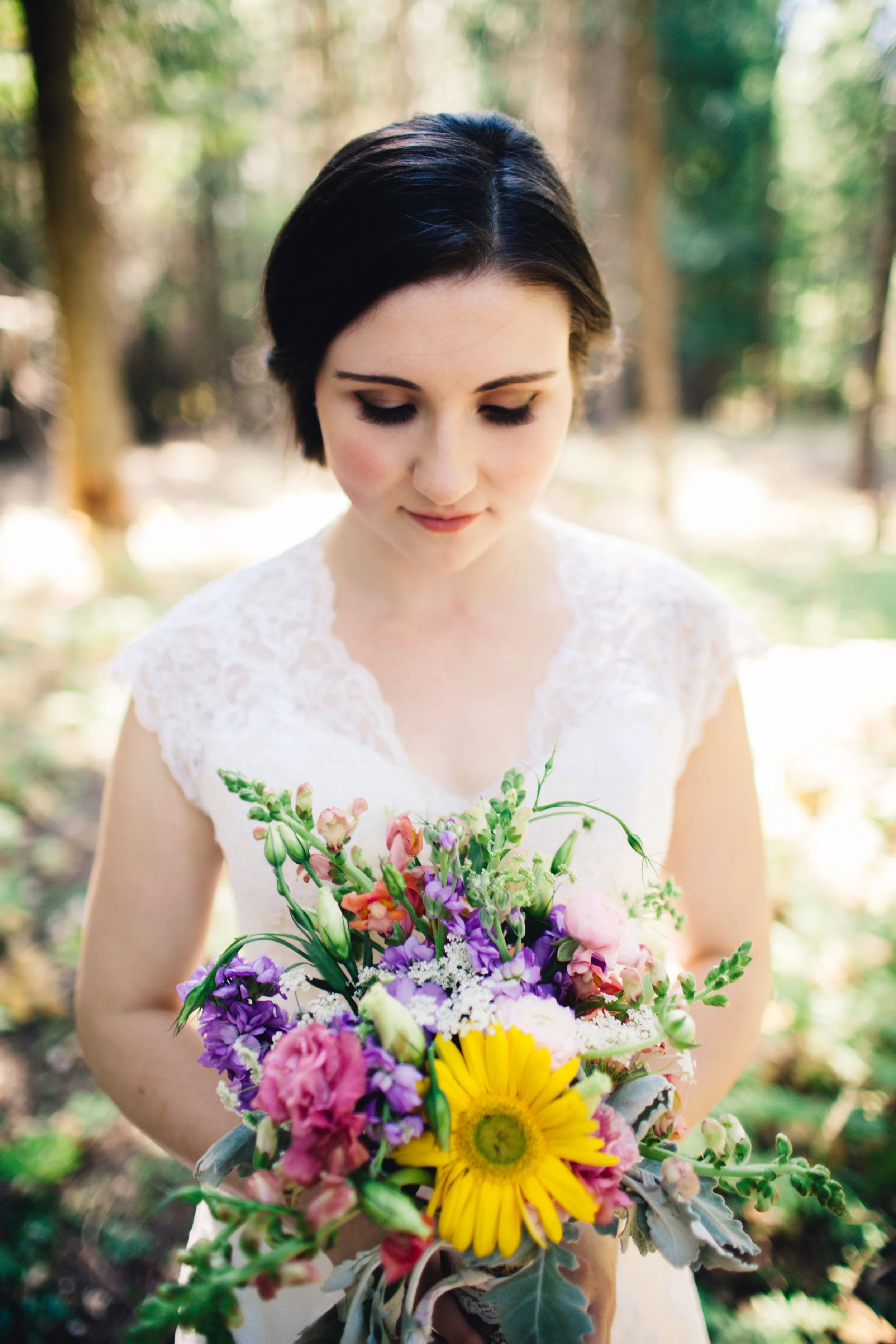 Brunette woman in white lace dress holding a colorful bouquet of flowers in a forest setting.