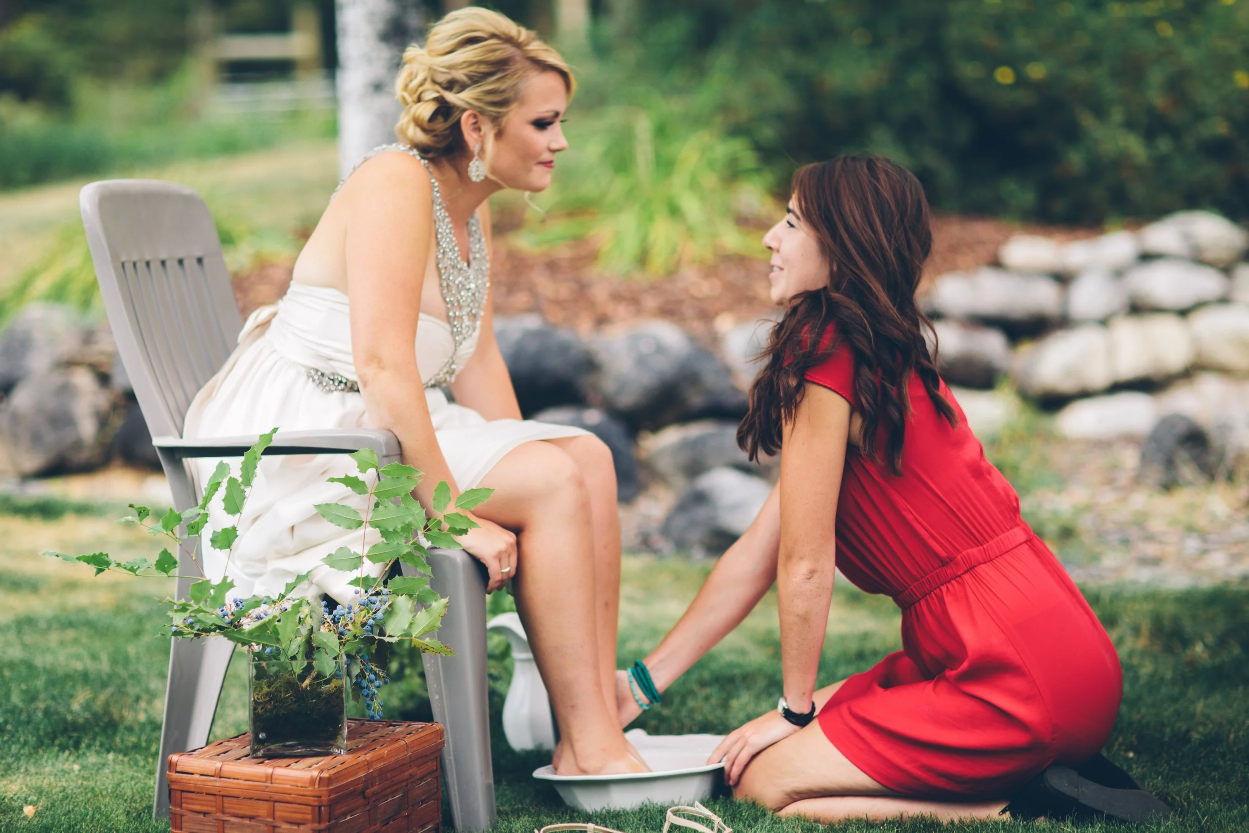 A bride sitting on a chair outdoors getting her feet washed by a woman kneeling on the grass, with a garden backdrop of rocks and trees.