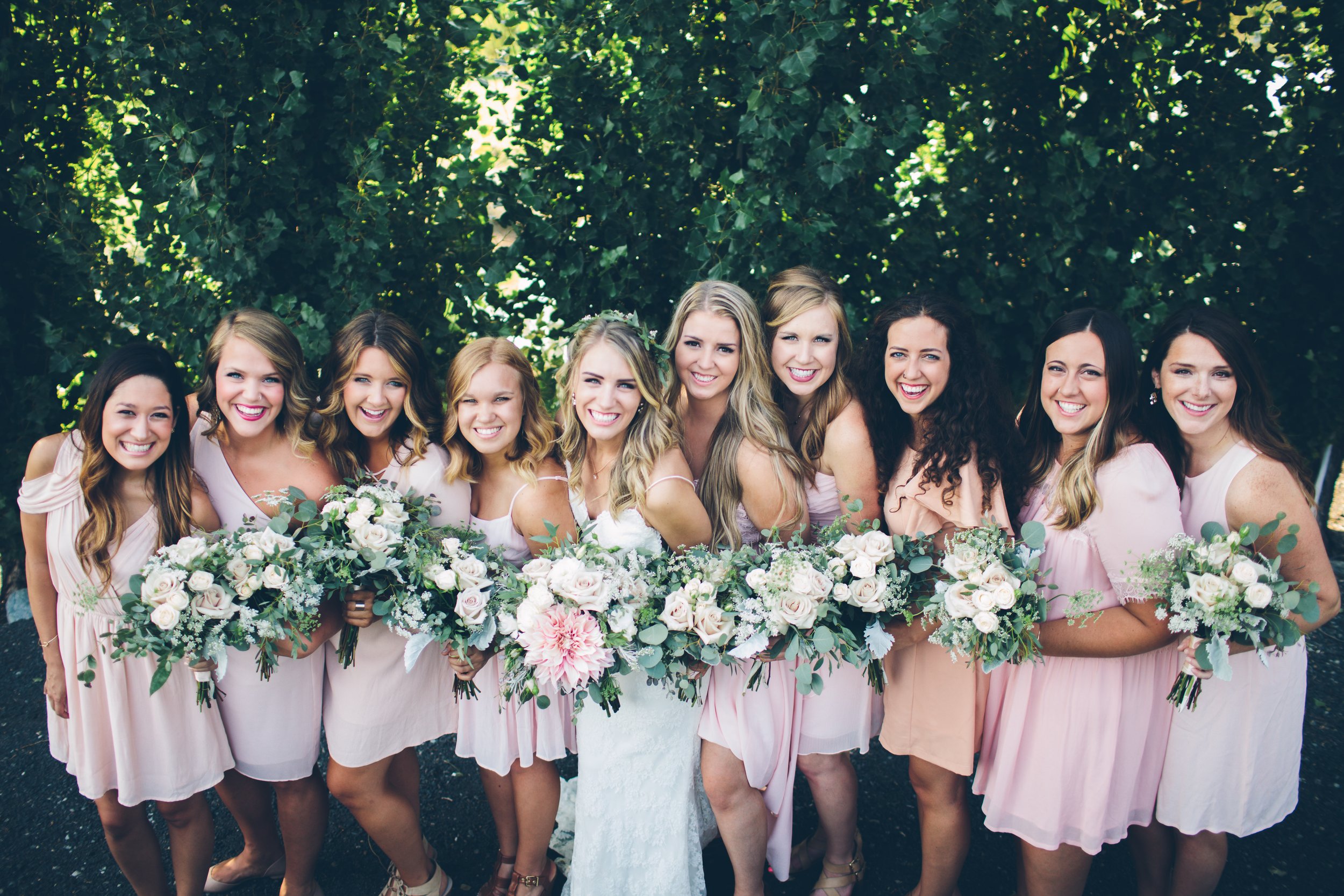 A group of eleven women dressed in light pink dresses, holding floral bouquets, standing outdoors in front of green foliage, smiling for a photo.