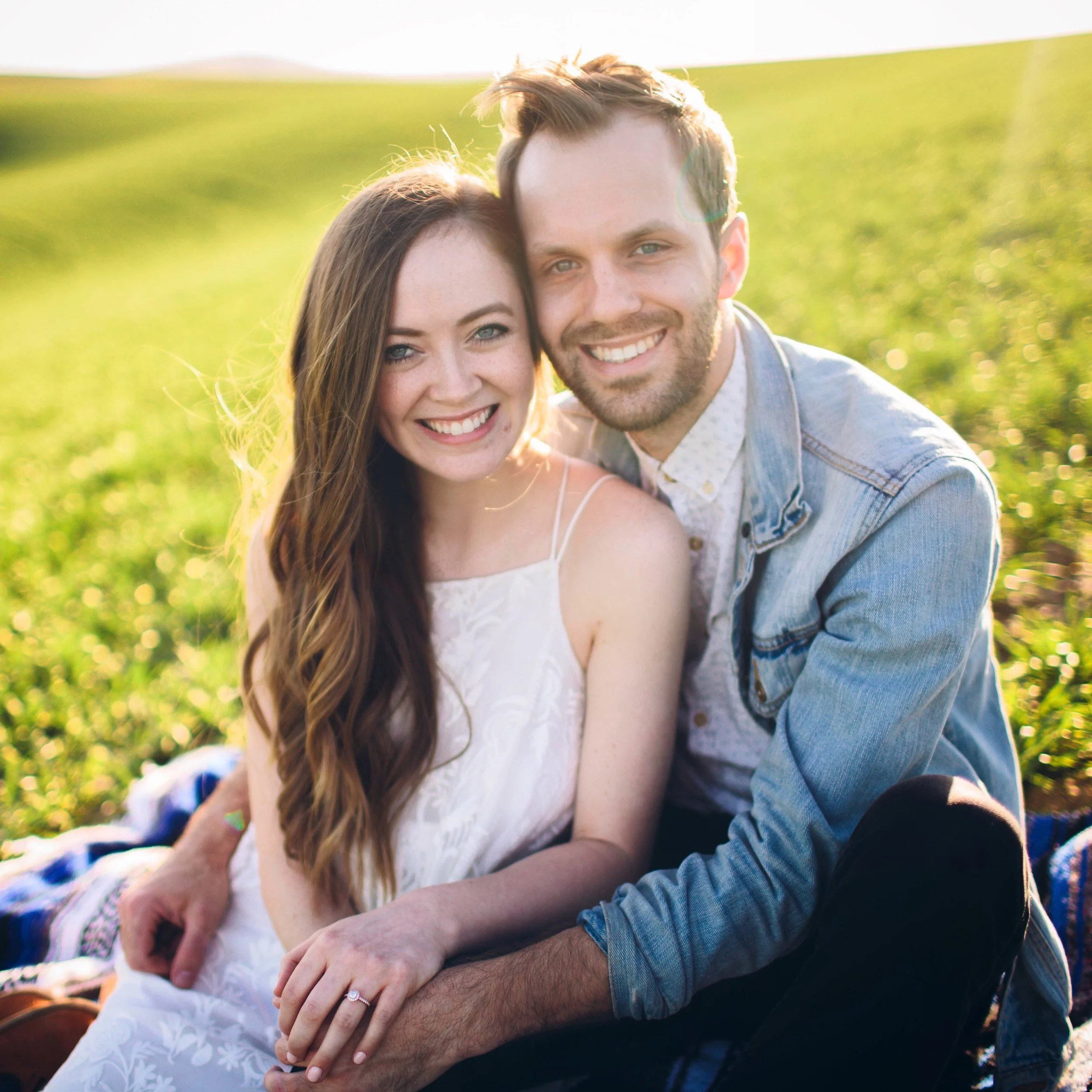 A young couple sitting on a blanket in a grassy field, smiling at the camera during sunset.