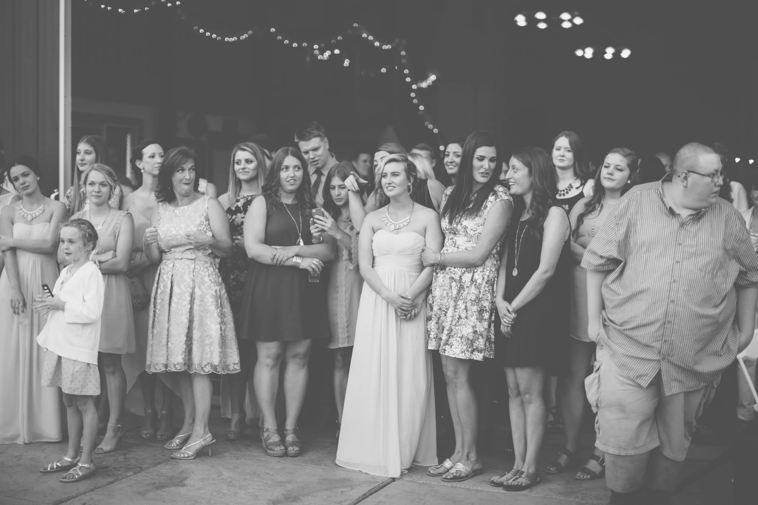 Group of women and a man at an indoor event, some wearing formal dresses, standing together, with string lights hanging overhead.
