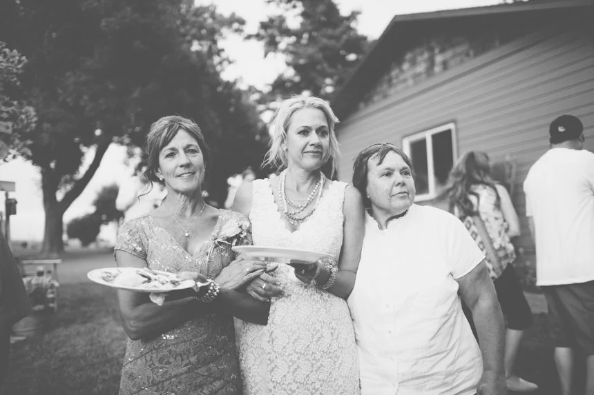 Three women standing outdoors at a gathering, with two holding plates of food, in front of a house and trees.