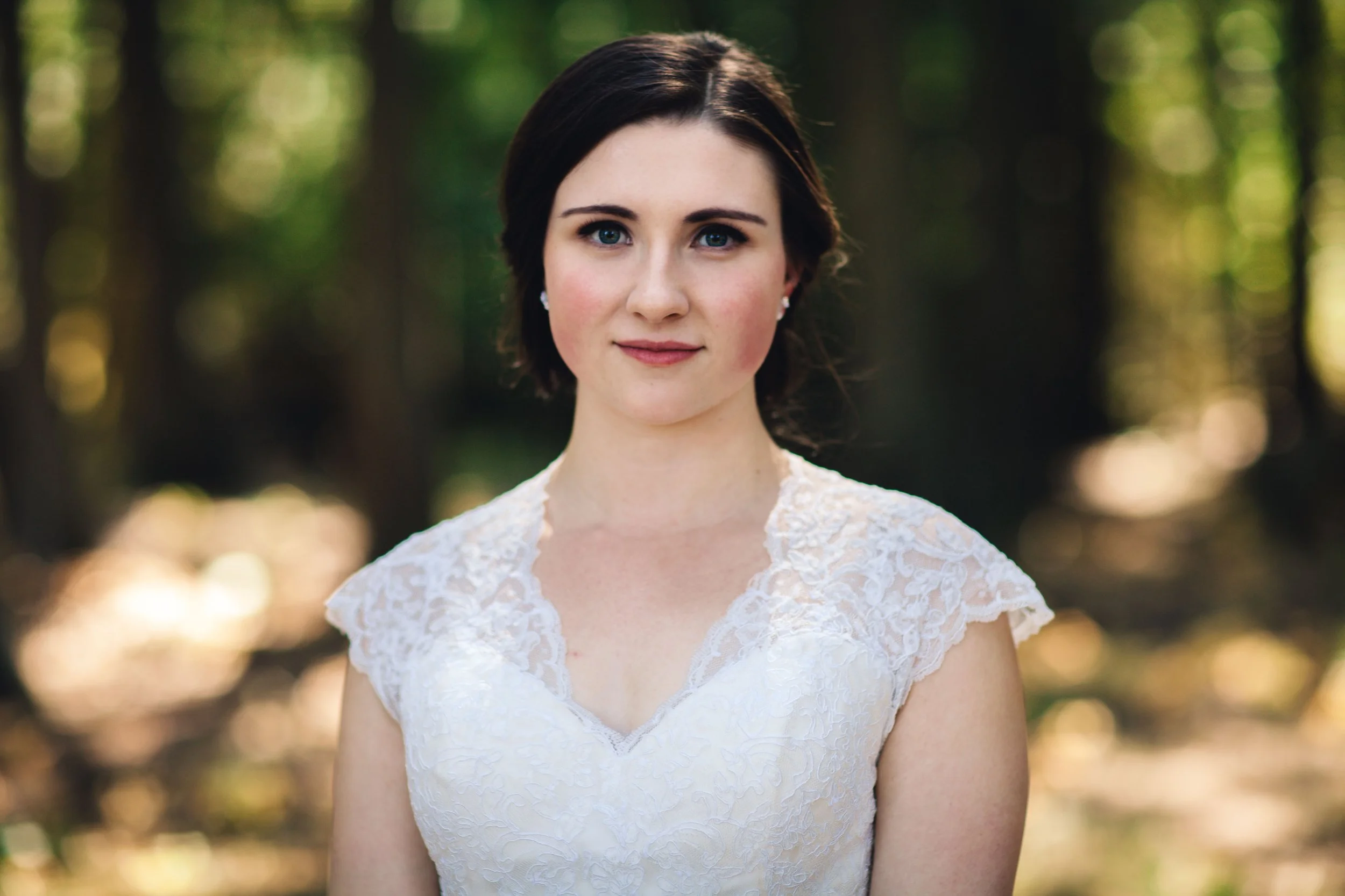 Portrait of a young woman with dark hair and blue eyes wearing a white lace dress, standing outdoors in a forest with sunlight filtering through trees.