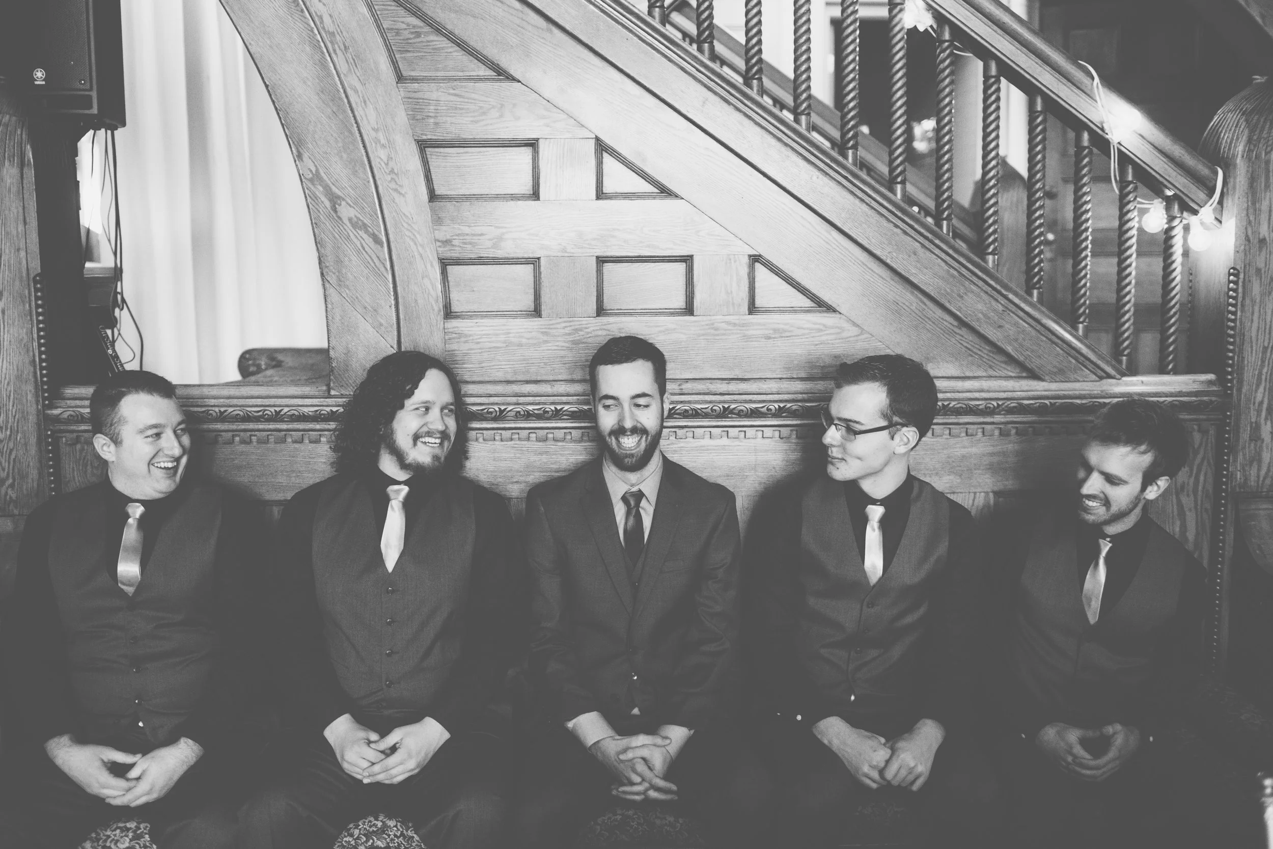 Five men in formal attire sitting in a row on a sofa, smiling and conversing, behind a wooden staircase.