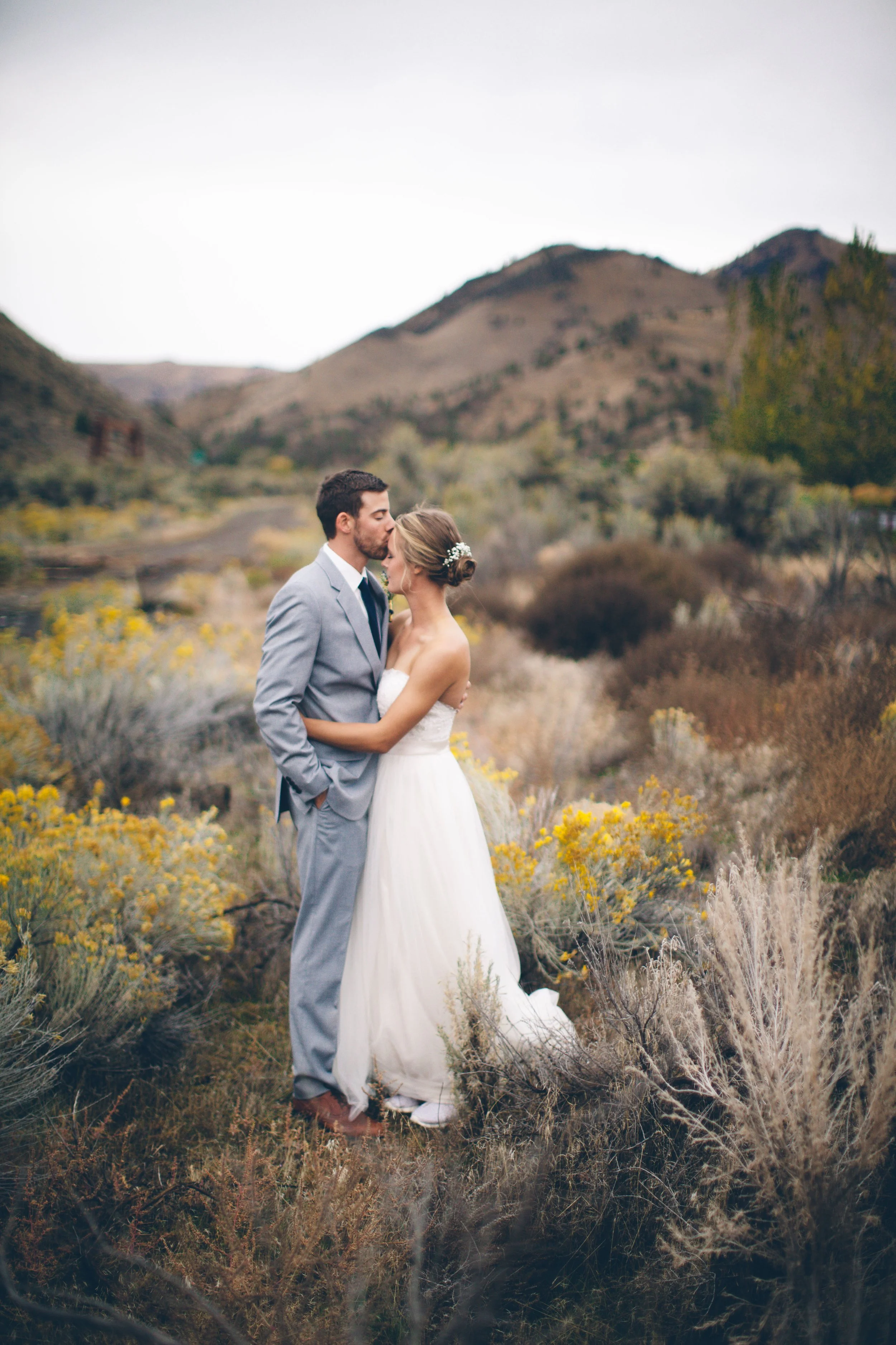 A bride and groom standing in a desert landscape with mountains in the background. The groom is kissing the bride on the forehead.