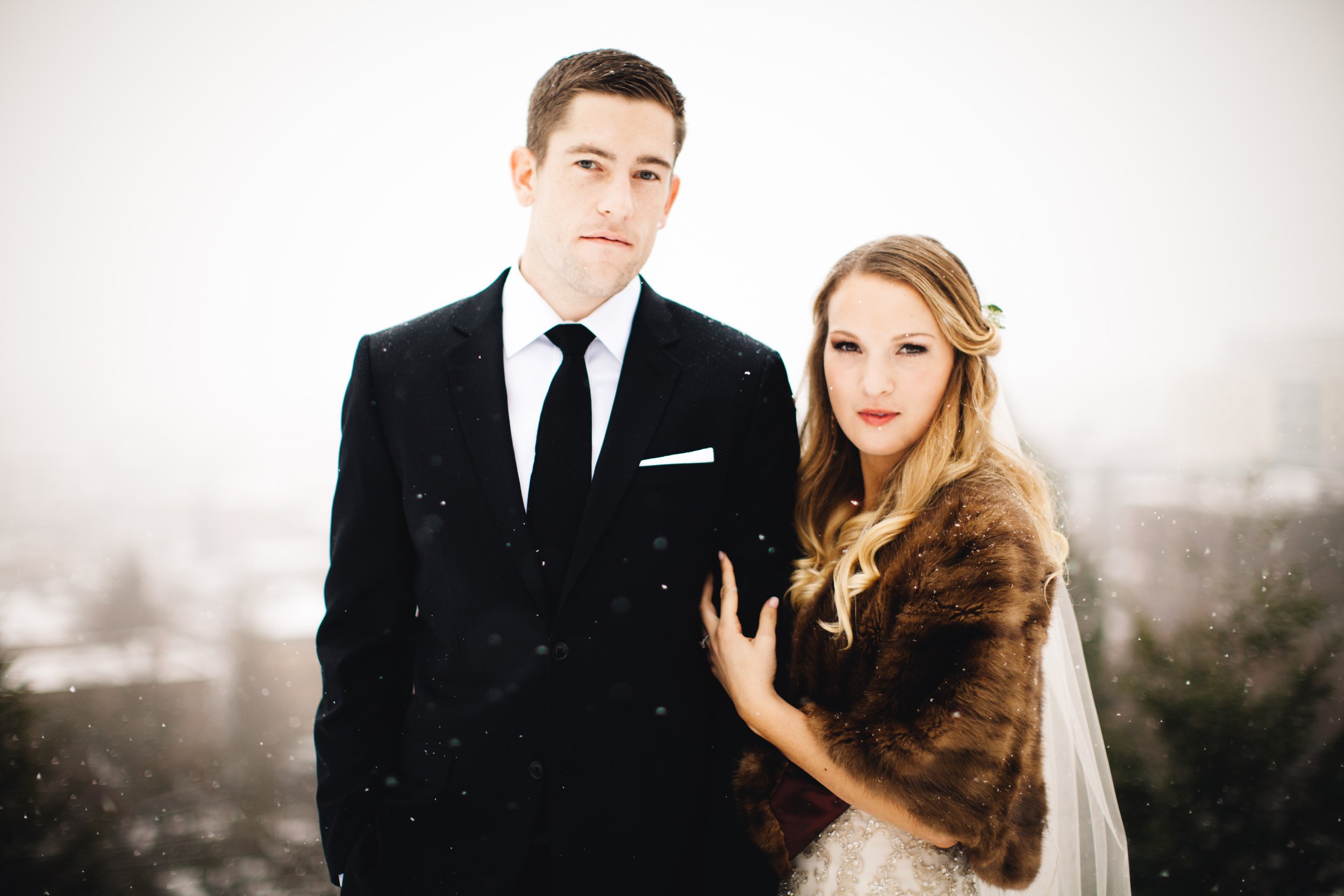 A man in a black suit and tie standing next to a woman with long blonde hair wearing a wedding gown and a brown fur stole outdoors in snowy weather.