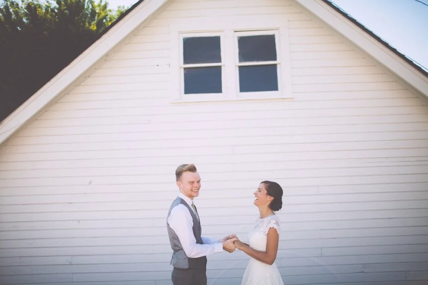 A newlywed couple holding hands and smiling in front of a white house with a gable roof and two small windows.