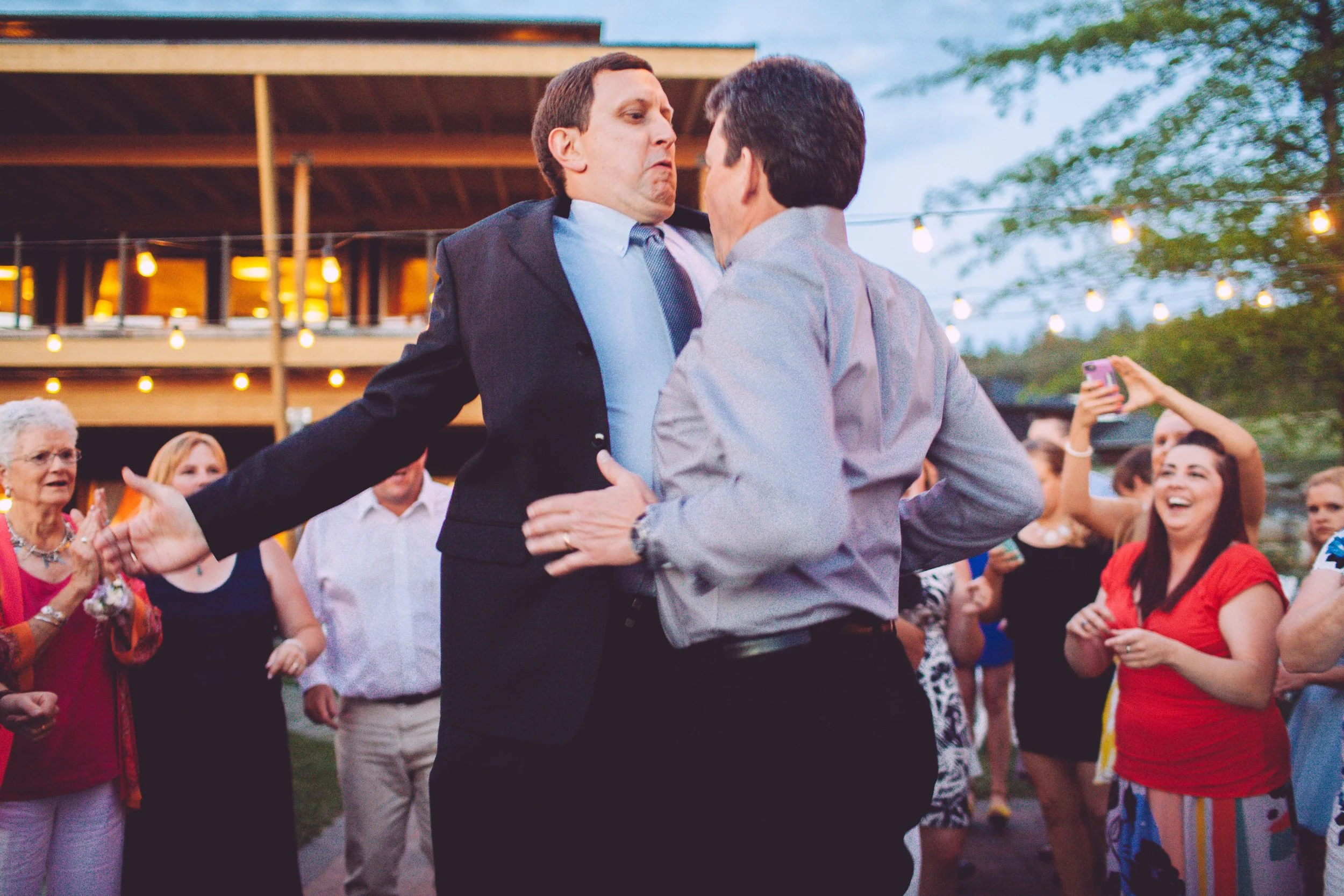Two men dancing closely at an outdoor event with string lights and people watching and cheering in the background.