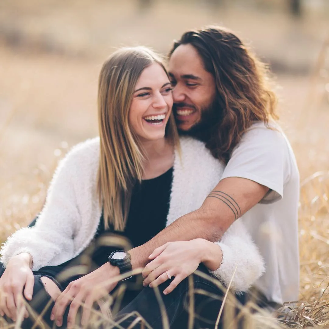 A smiling couple sitting close together in a field of tall, golden grass.