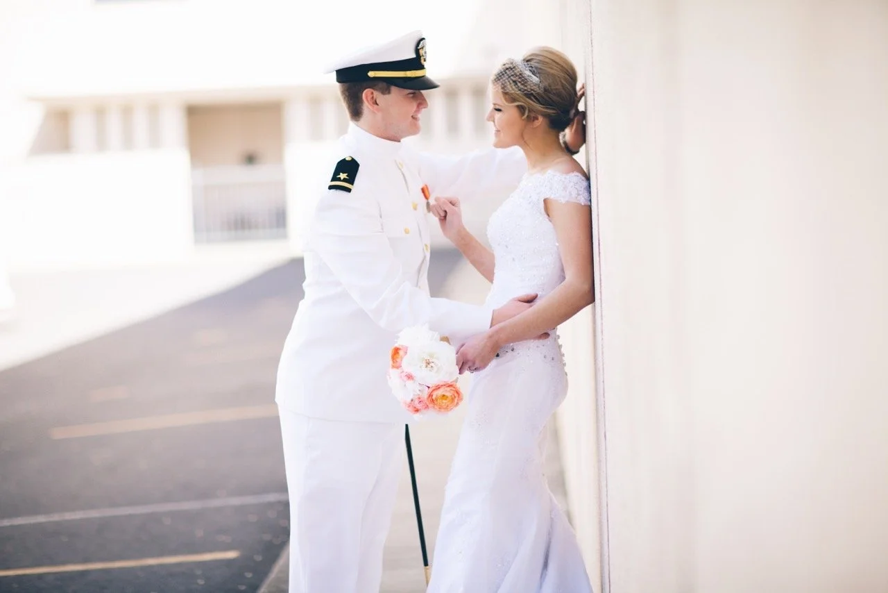A bride and groom in wedding attire sharing a moment outdoors, with the groom in a military uniform and the bride in a white lace wedding dress, holding a bouquet of flowers.
