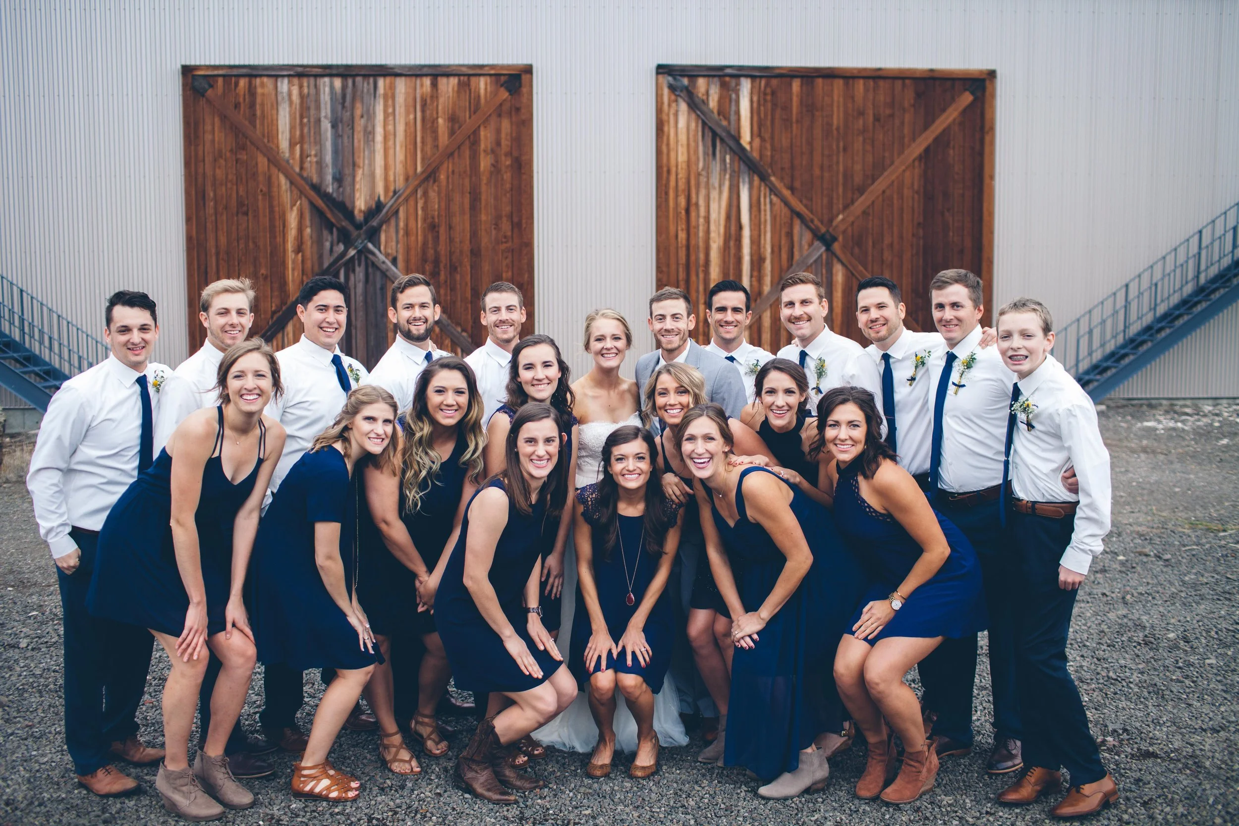 A group of people dressed in semi-formal attire, posing outdoors in front of a large wooden barn with sliding doors and metal siding, during a celebration or wedding.