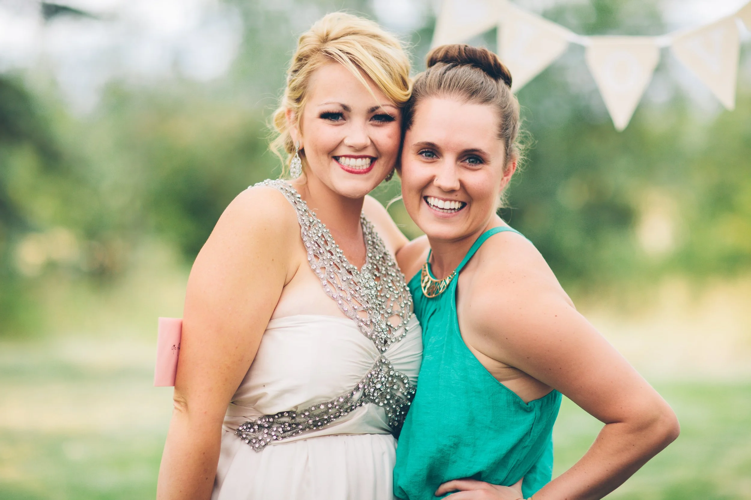 Two women smiling outdoors at a celebration, one wearing a white dress with sparkling embellishments, and the other in a teal sleeveless dress.