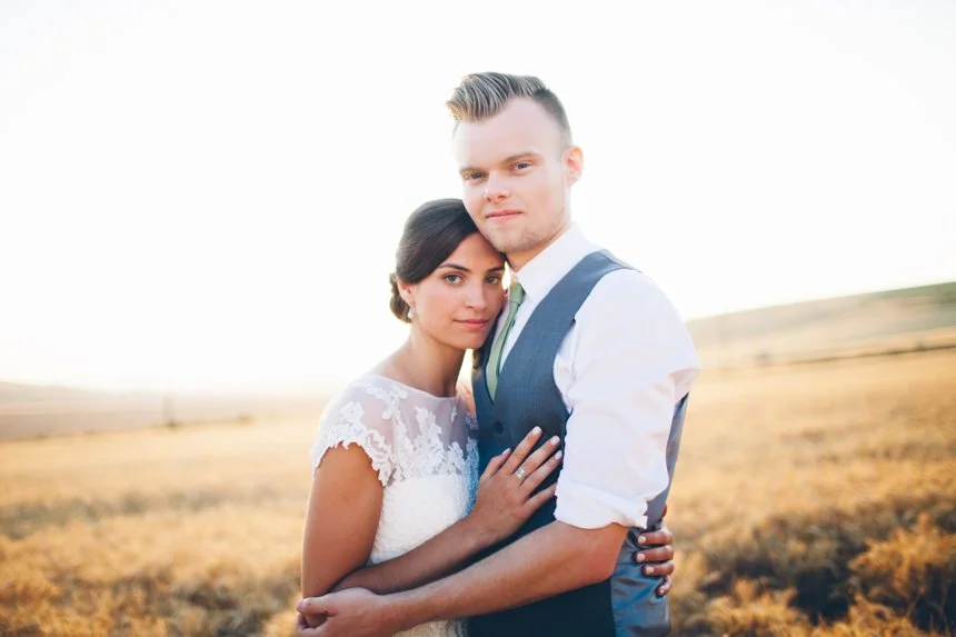 A couple embraces outdoors during sunset, with the woman in a lace dress and the man in a vest and rolled-up sleeves, standing in a golden field.