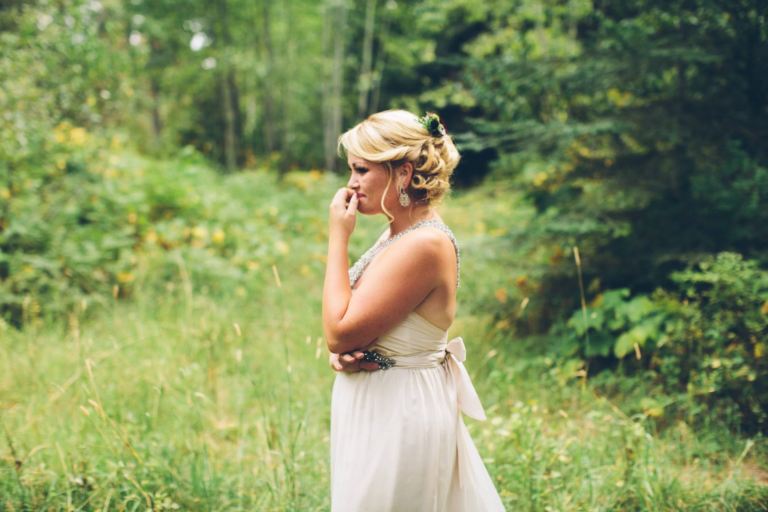 A bride in a white wedding dress standing in a green forest, holding her hand to her face in a contemplative pose.