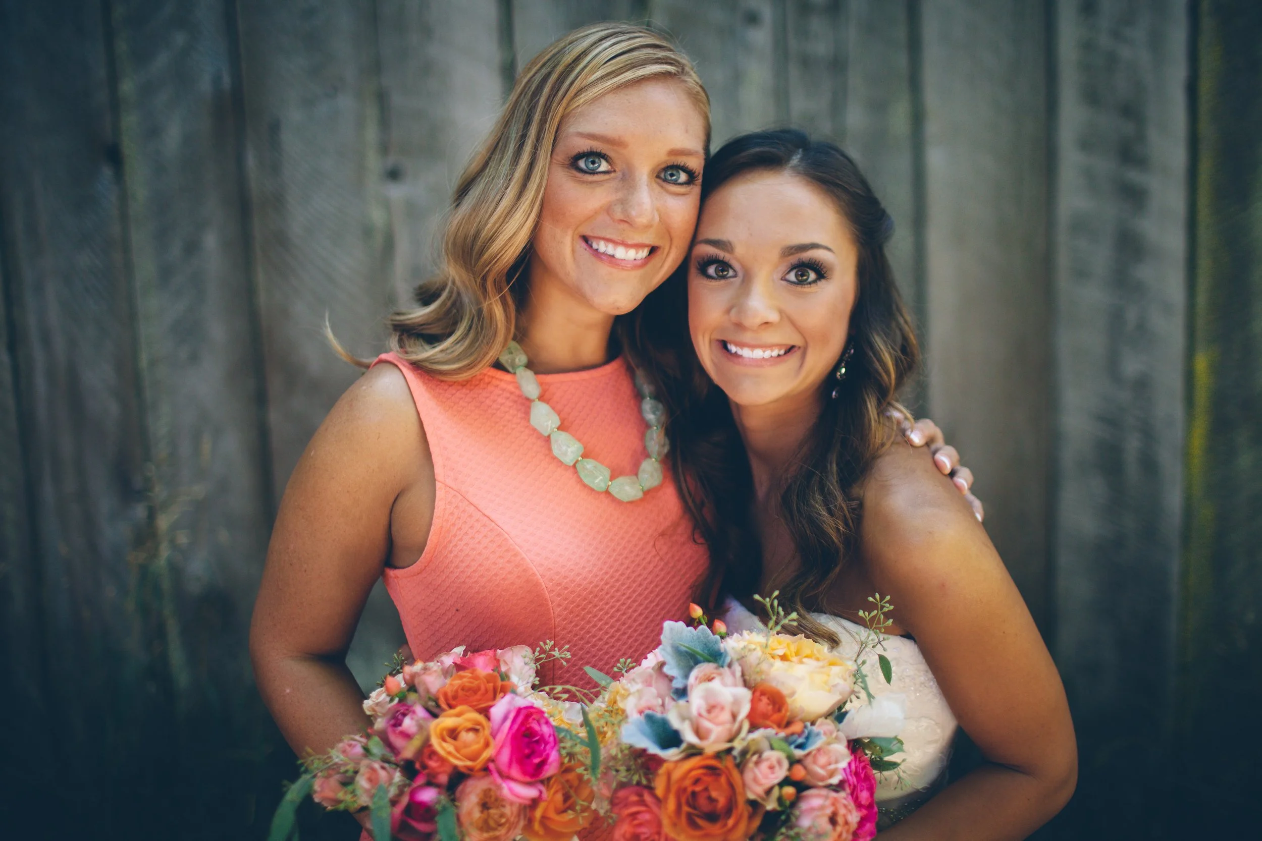 Two smiling women, one in a coral dress and the other in a white dress, standing close together outdoors with a wooden fence behind them, holding colorful bouquets of flowers.