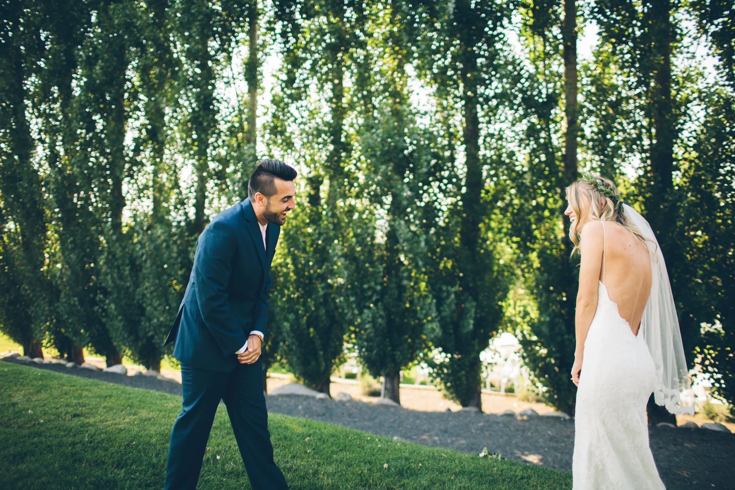 A groom in a navy suit is smiling and bowing as he looks at a bride in a white wedding dress and veil, standing outdoors in a lush green park with tall trees in the background.