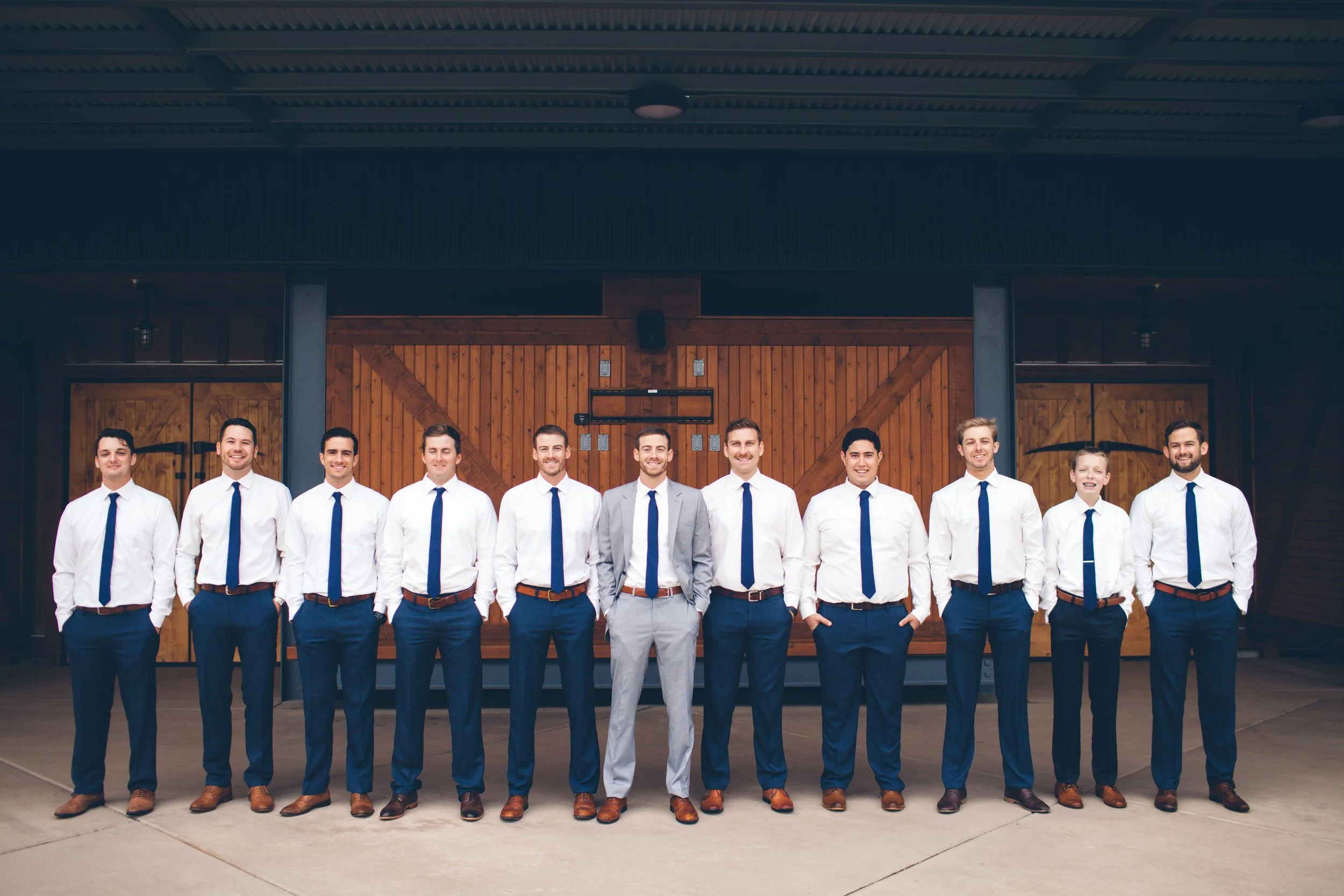 A group of eleven men dressed in white shirts, navy ties, and navy pants, standing outdoors in front of a wooden barn door, with one man in a gray suit at the center.