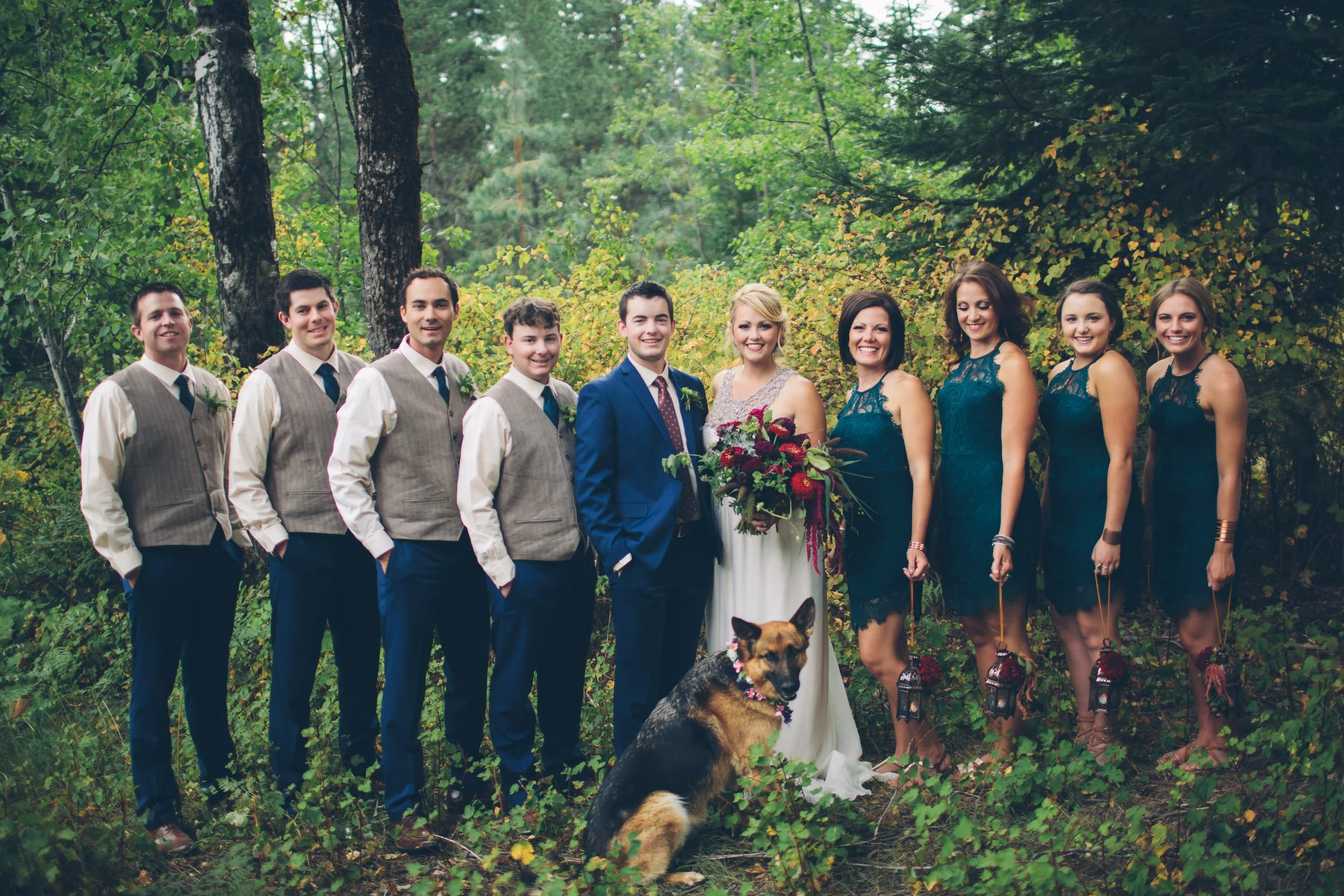 A wedding party standing in a forest, including four men in vests and ties, a bride holding a large bouquet, a groom in a blue suit, and four women in matching teal dresses, with a dog sitting in front of them.