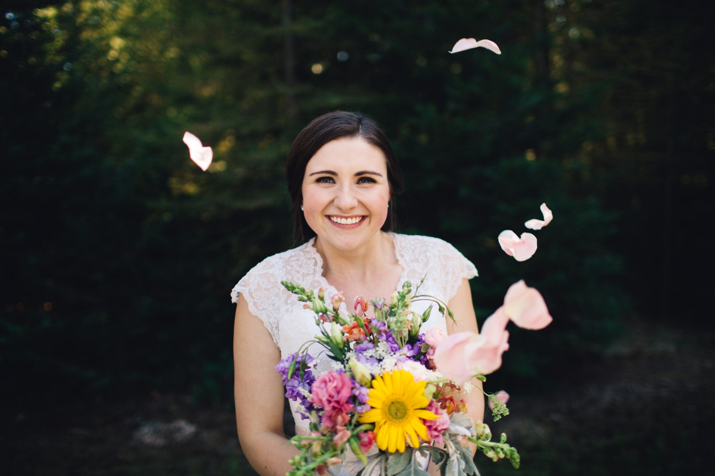 A woman smiling and holding a colorful bouquet of flowers outdoors, with petals falling around her.
