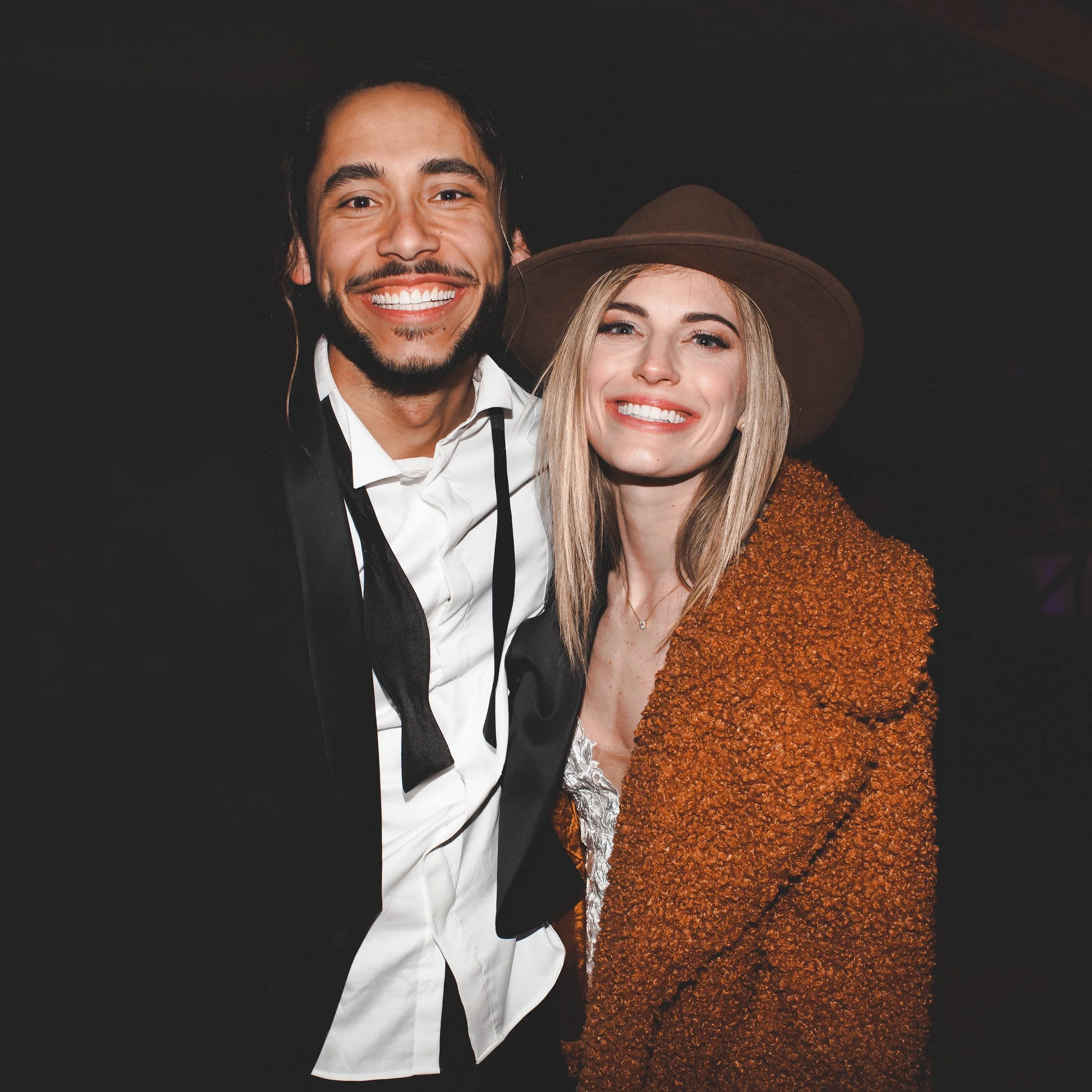 A smiling man and woman standing close together indoors, with the man wearing a black suit and the woman wearing a brown hat and a textured orange-brown coat.