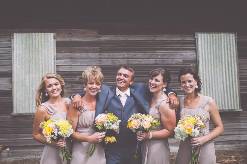 Group of five people, four women and one man, dressed in formal attire, smiling and holding bouquets of flowers, standing outdoors in front of a rustic wooden building.