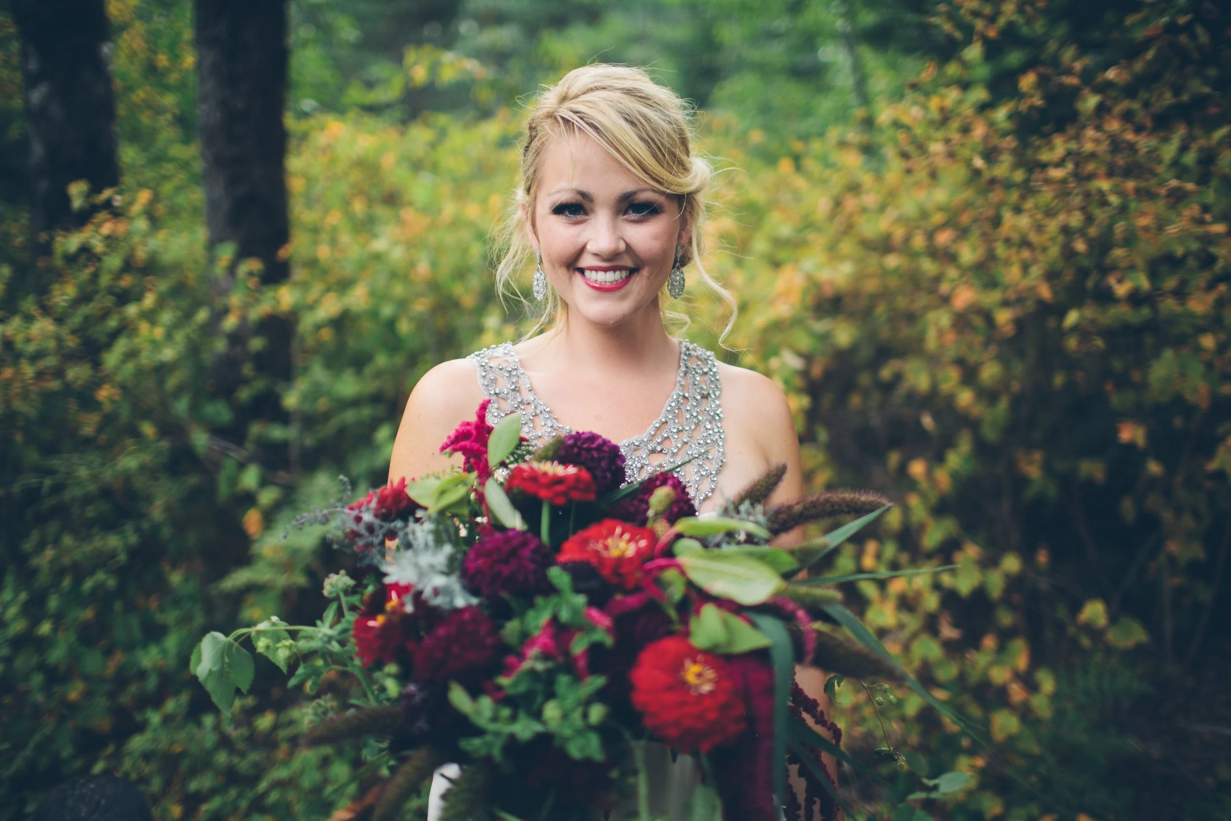 A woman in a wedding dress holding a large bouquet of flowers outdoors with green trees and foliage in the background.