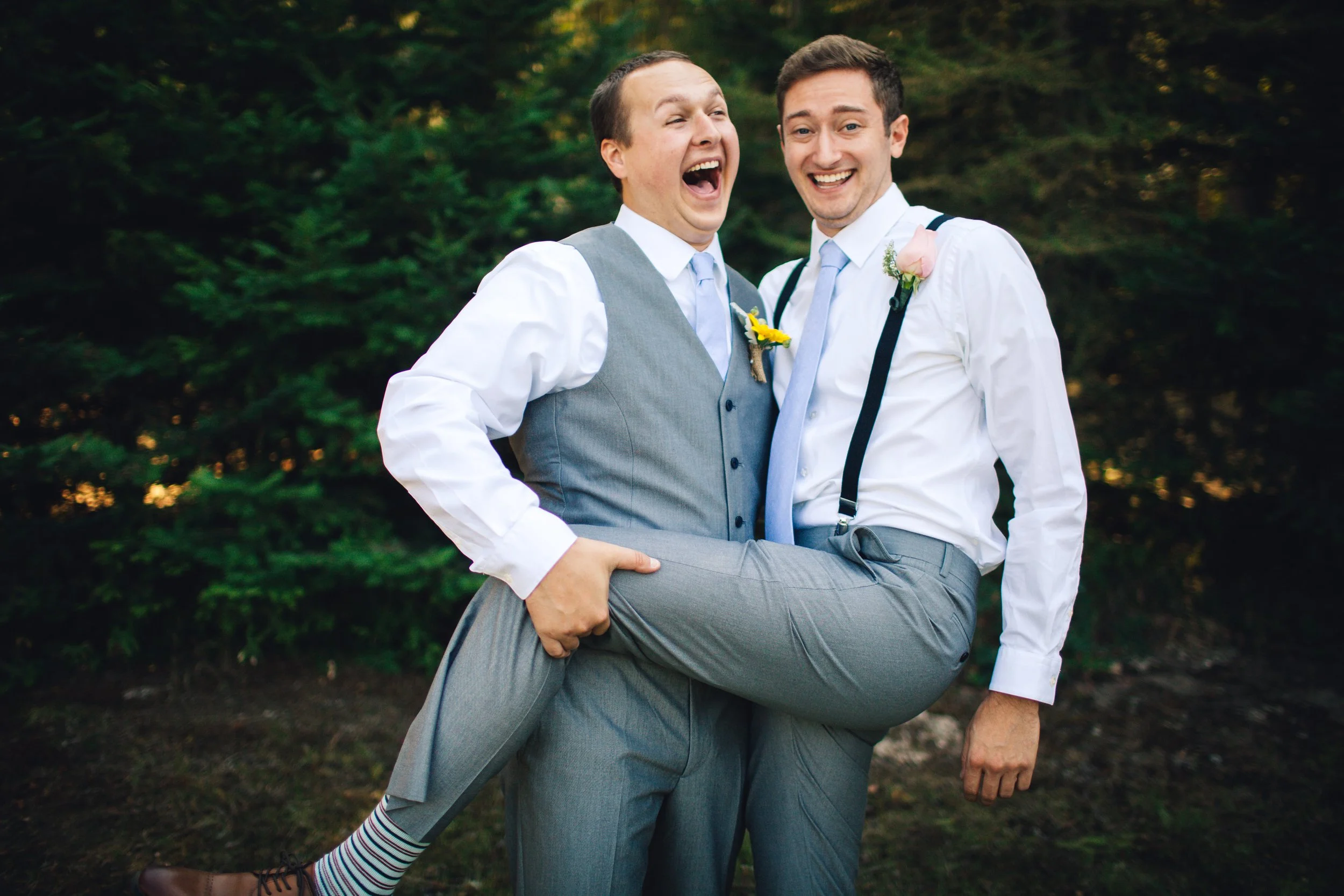 Two men in formal attire, smiling and laughing, with one lifting the other in the outdoors with trees in the background.