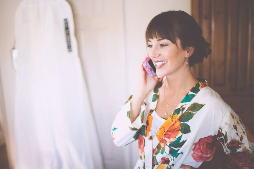 A woman smiling and talking on a pink cellphone, wearing a white floral jacket, indoors with wooden cabinets.