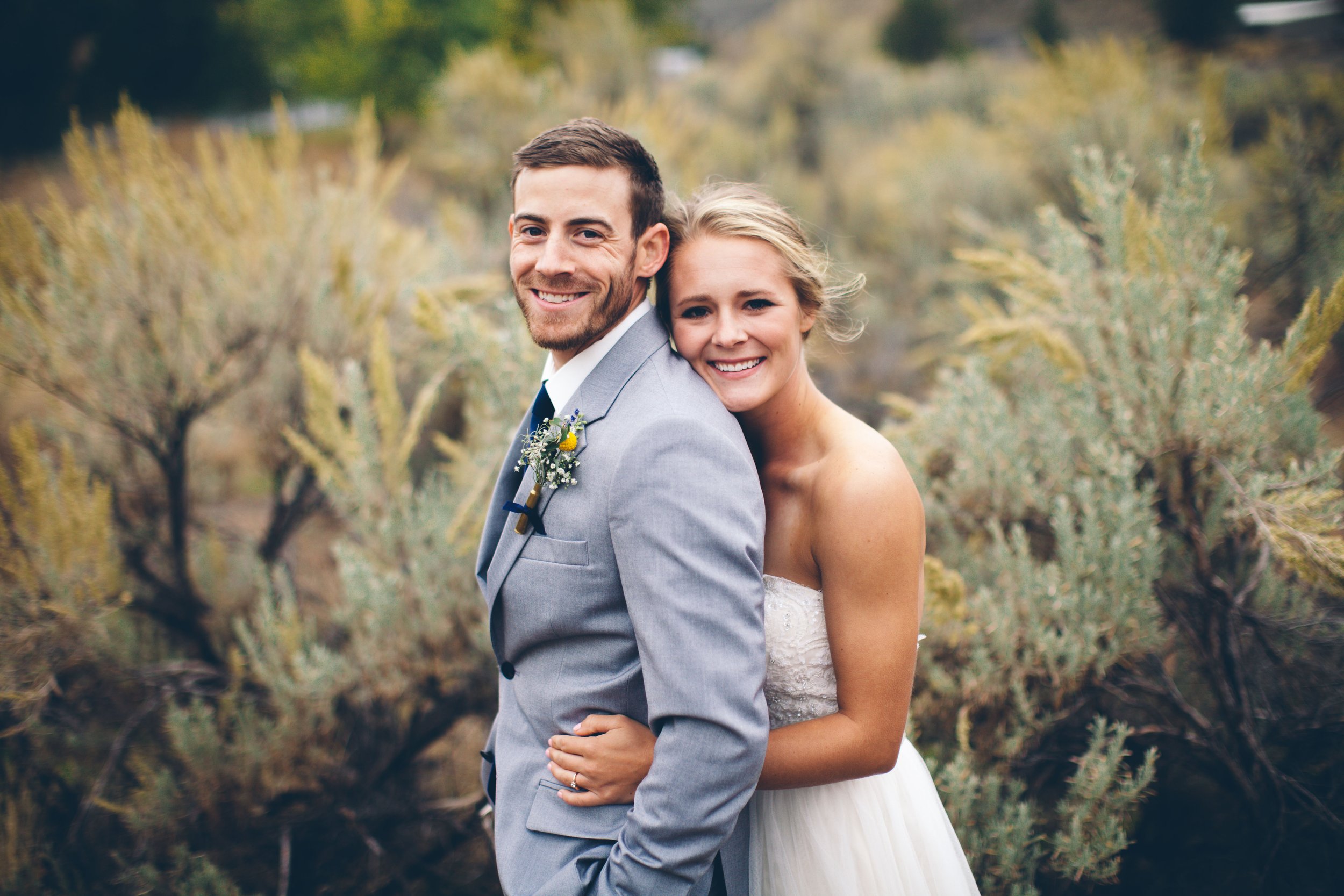 A smiling bride and groom outdoors with yellow and green foliage in the background. The bride, with blonde hair, is hugging the groom from behind. The groom, with brown hair and a beard, is wearing a gray suit with a boutonniere, and the bride is wea
