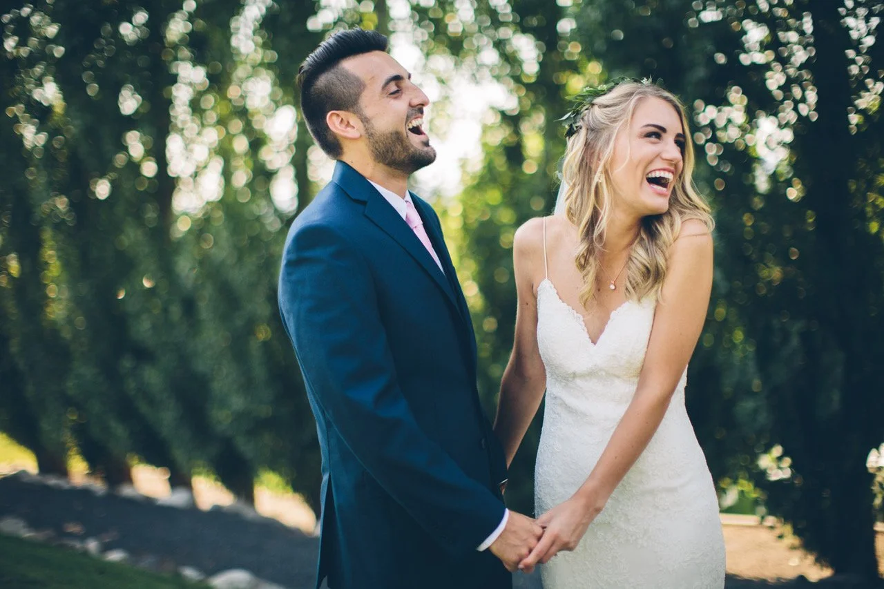 A smiling couple dressed in wedding attire, holding hands outdoors with trees in the background.
