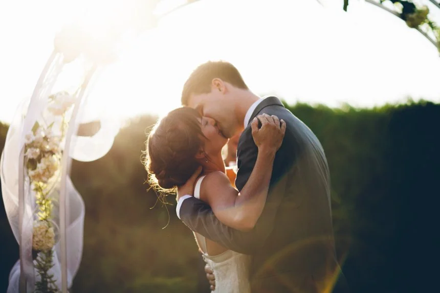 A bride and groom sharing a kiss outdoors during sunset, with a wedding arch decorated with flowers in the background.