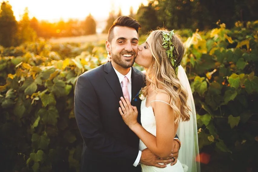 A newlywed couple sharing a kiss in a vineyard at sunset, with lush green vines in the background.