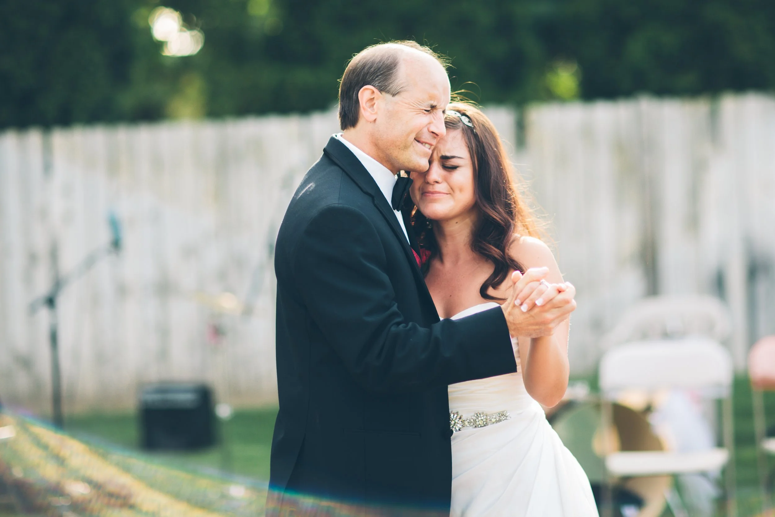 A bride and groom share an emotional dance, holding hands and closing their eyes, outdoors with a wooden fence and chairs in the background.