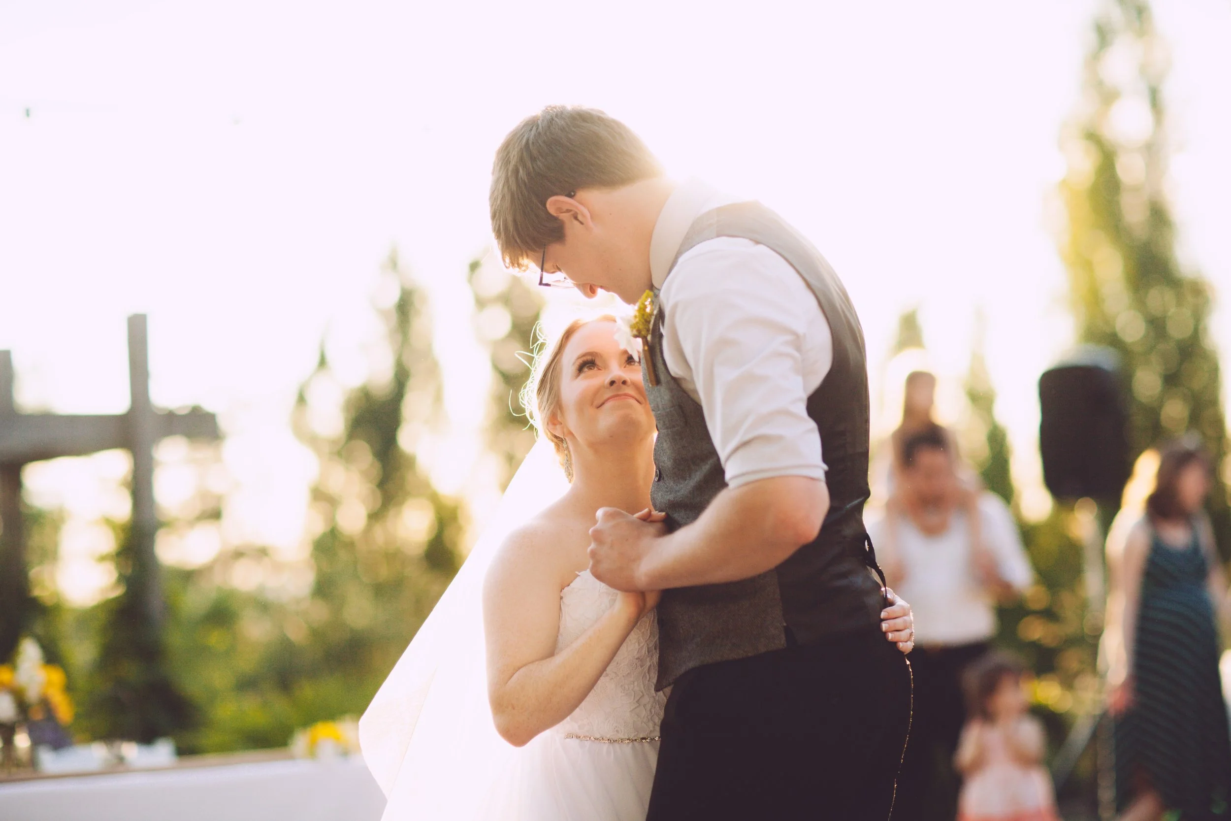 A bride and groom sharing a dance outdoors at sunset, with the bride gazing up at the groom, who is smiling down at her, surrounded by wedding guests.