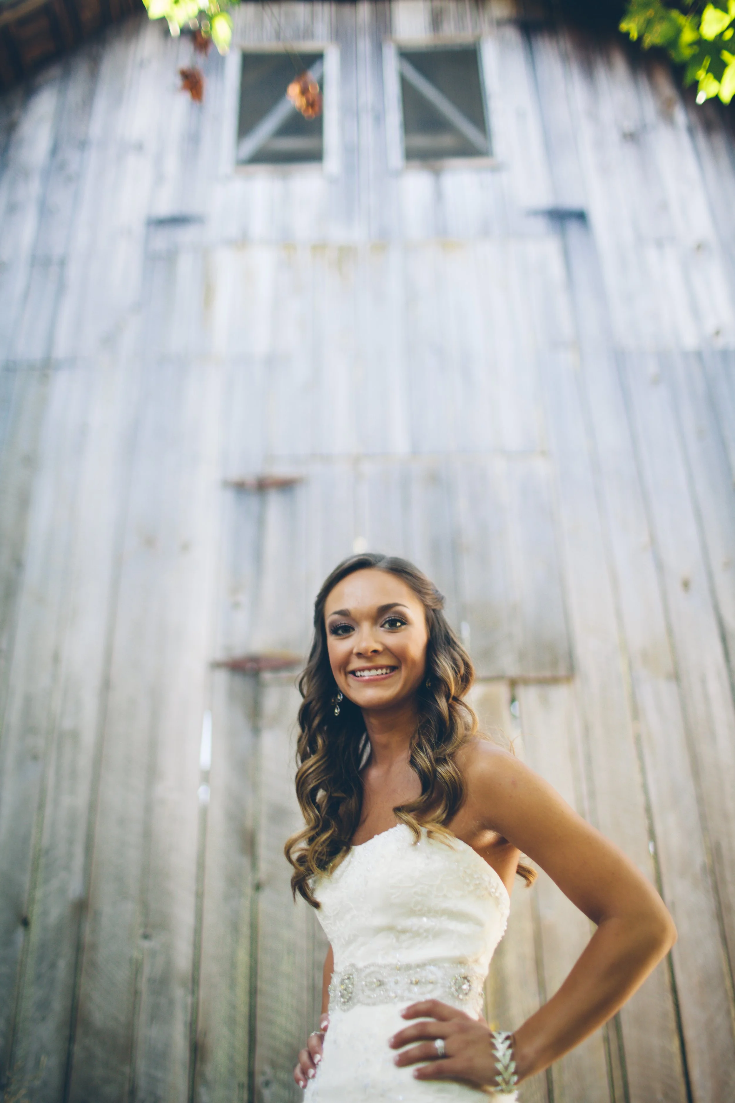 A smiling woman in a white wedding dress with long wavy hair, standing outdoors in front of a weathered wooden barn.