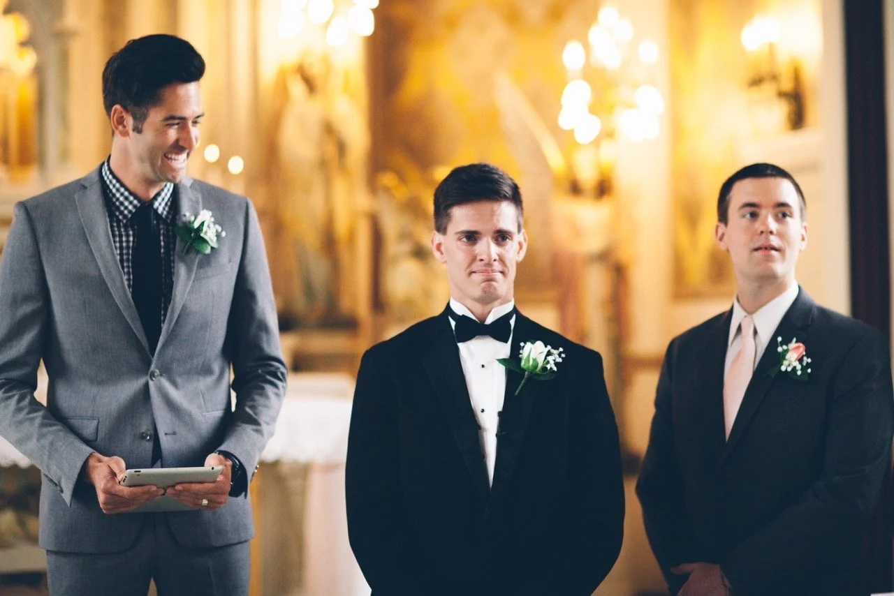 Three men dressed in formal tuxedos and suits participating in a wedding ceremony, with elaborate golden interior and sconces in the background.