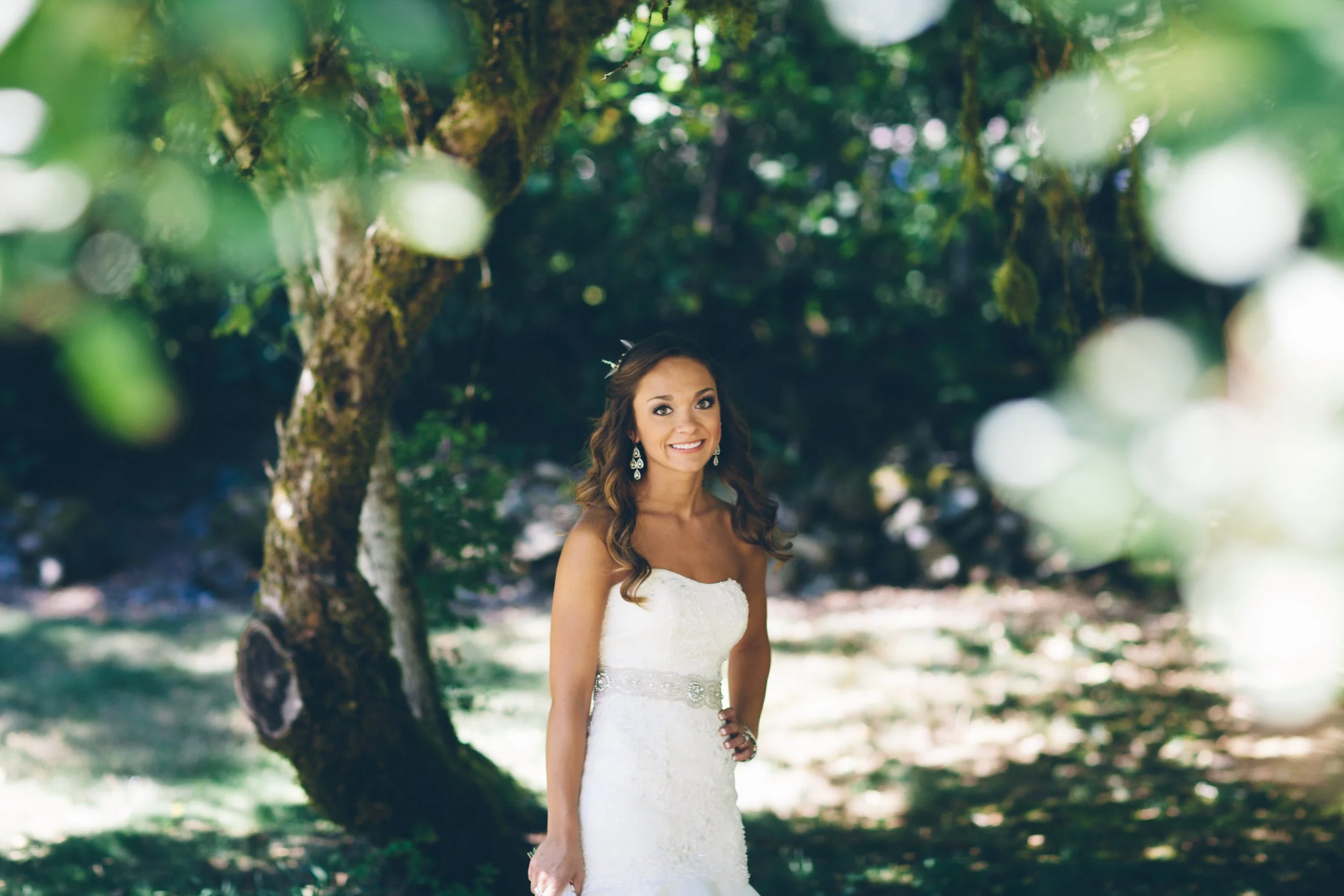 A smiling woman in a white wedding dress standing outdoors under a tree with green leaves.