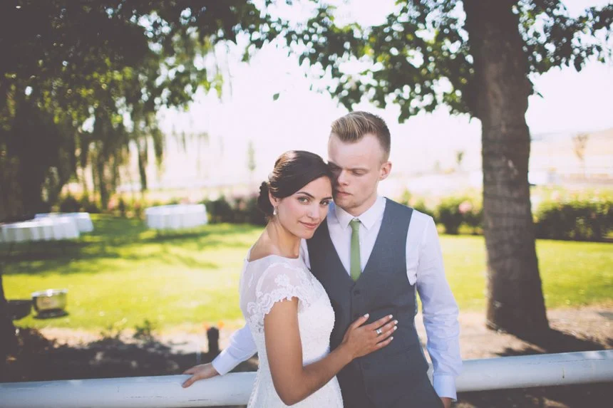 A newlywed couple outdoors on a sunny day, surrounded by trees and greenery, with a woman in a white lace wedding dress and a man in a gray vest and white shirt with a green tie.