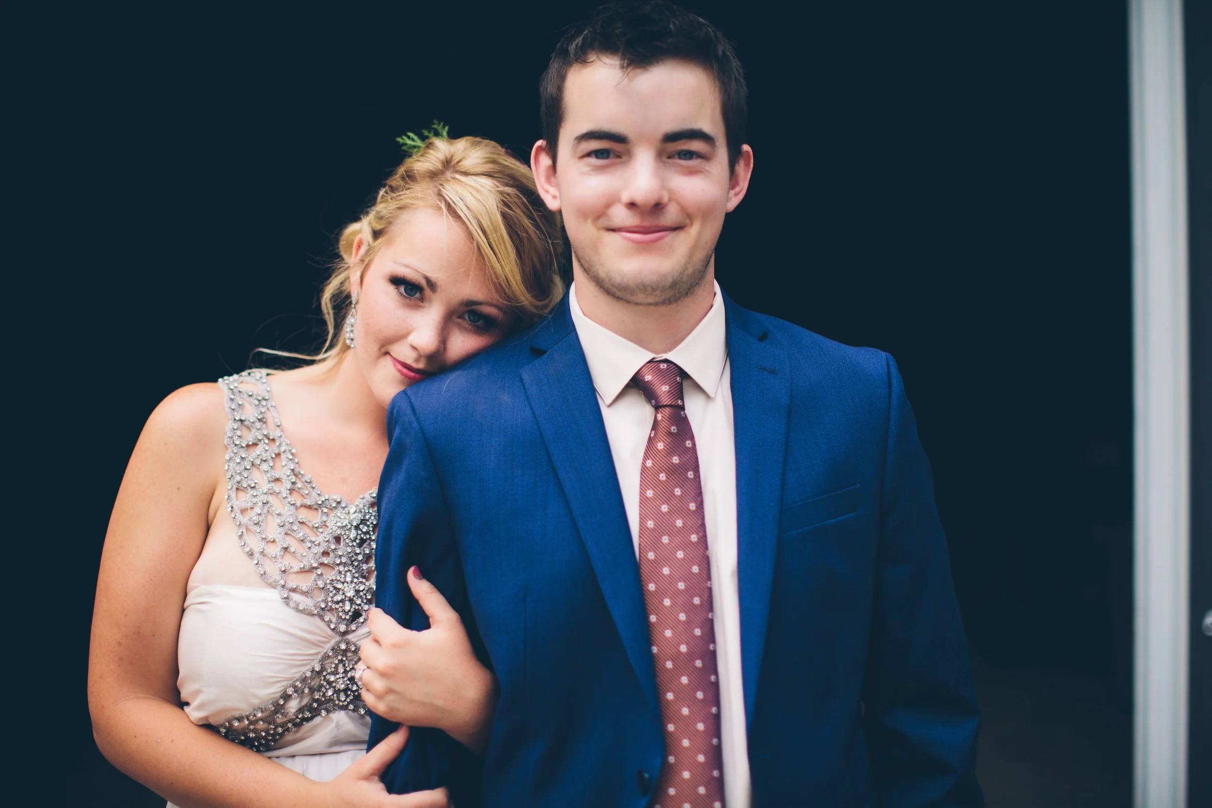 A young woman with blonde hair and a man with dark hair, both in formal attire, standing close together against a black background. The woman is leaning her head on the man's shoulder, smiling softly, while the man is smiling with a slight grin.