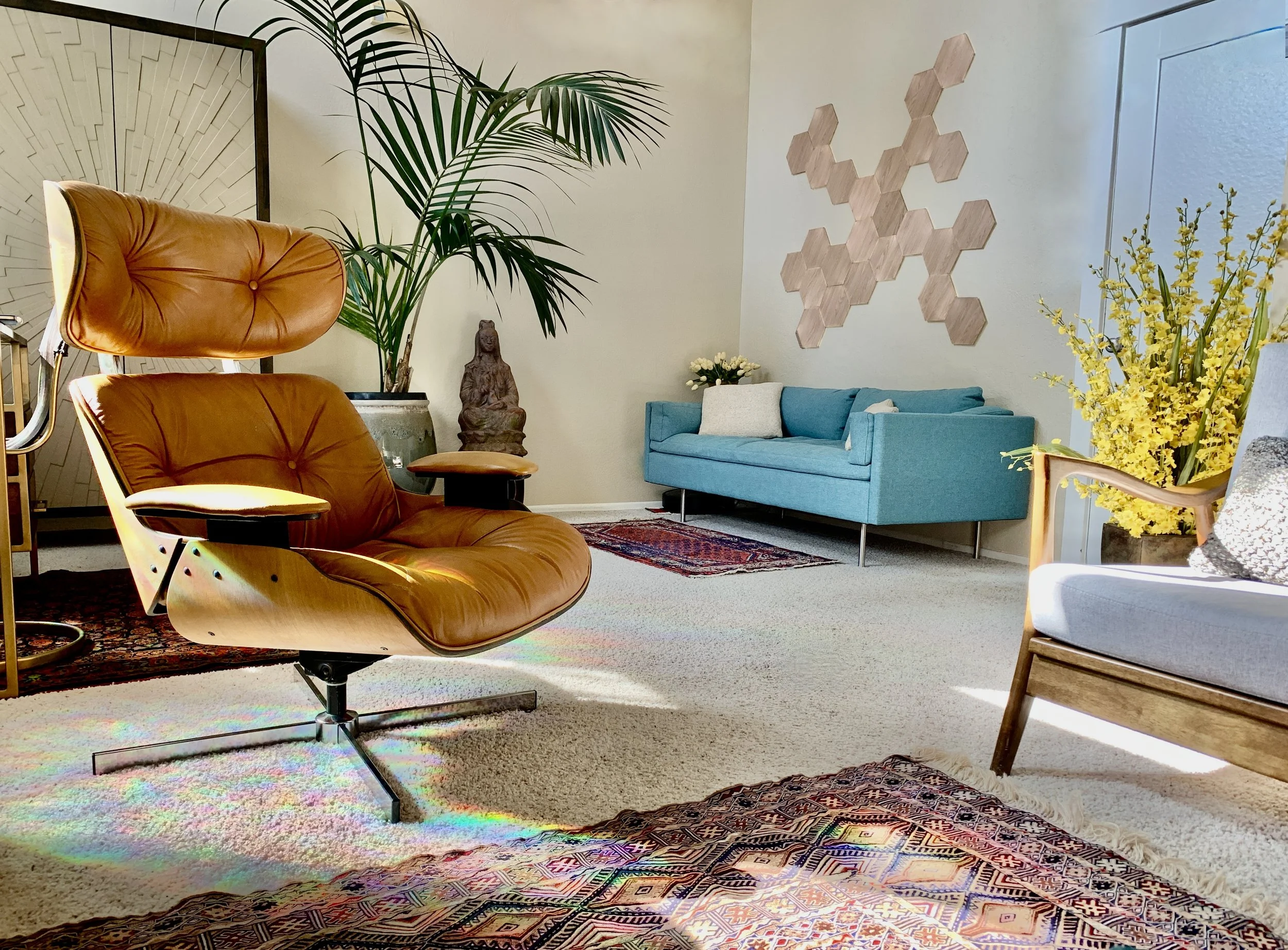 A modern living room with a tan leather armchair, a blue sofa, a potted plant, a Buddha statue, yellow flowers, a patterned rug, and wooden wall art.