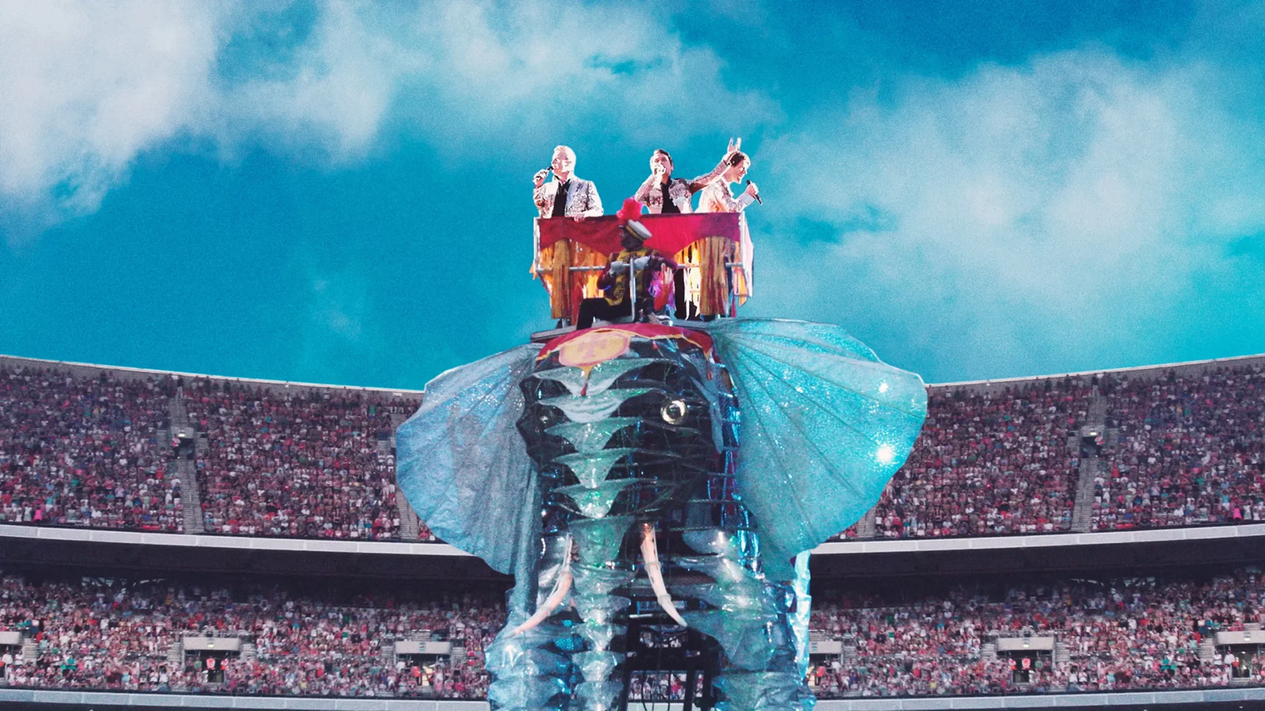 Performance on a large elephant-shaped float with performers and a music band, in a stadium filled with audience.