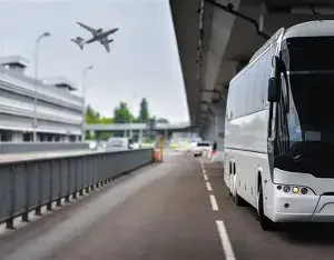 White bus parked on the side of a road near a bridge with an airplane flying overhead in the sky.