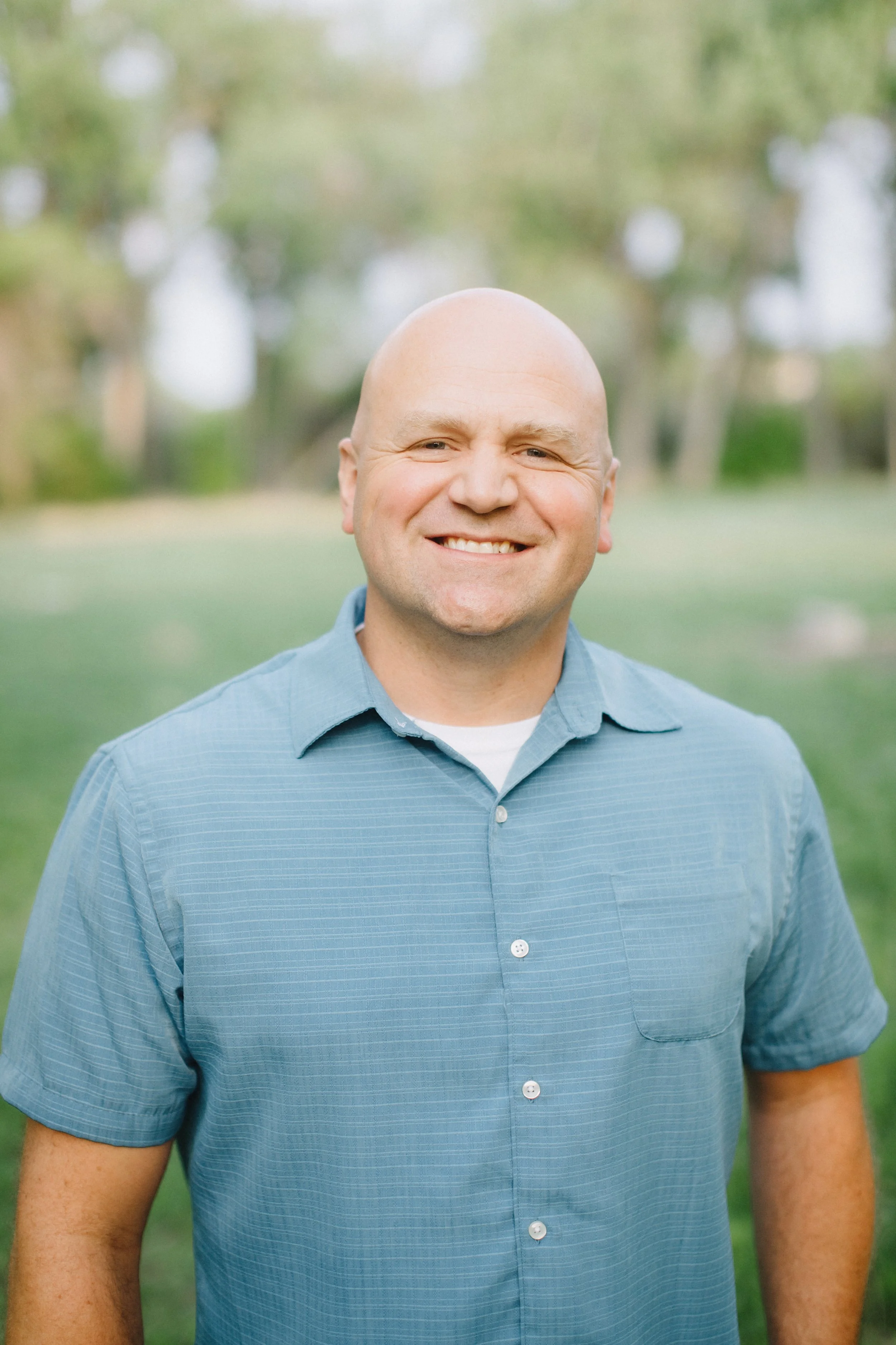 Smiling man wearing a blue short-sleeve button-up shirt, standing outdoors with blurred green trees and grass in the background.