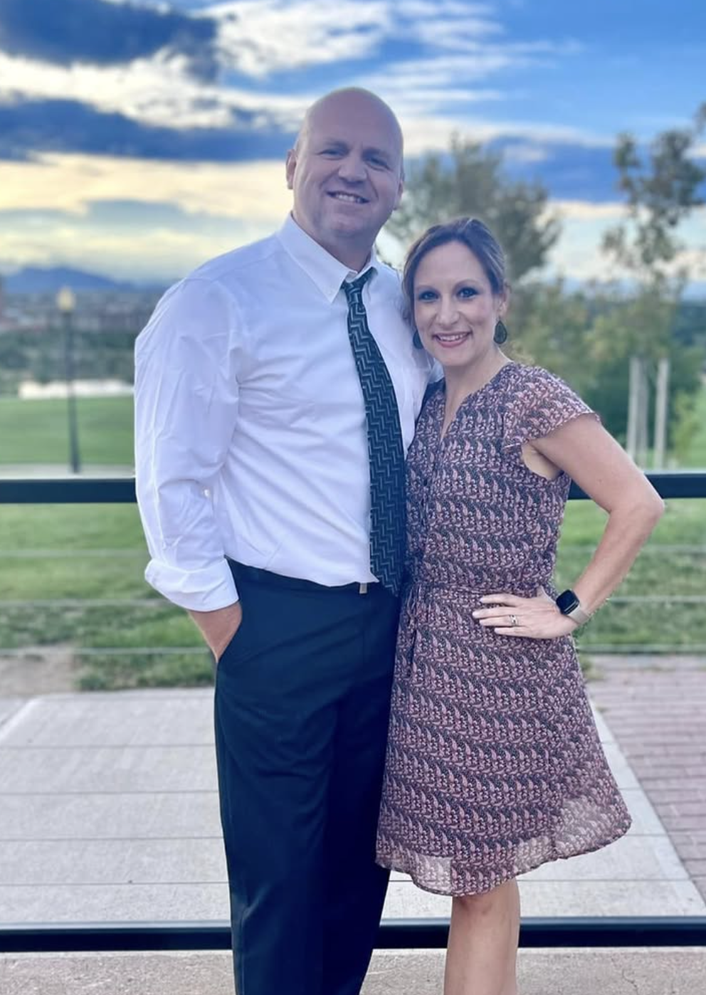 A smiling man and woman standing outdoors, with the man in a white shirt and tie and the woman in a patterned dress, against a scenic background with trees, a river, and a cloudy sky.